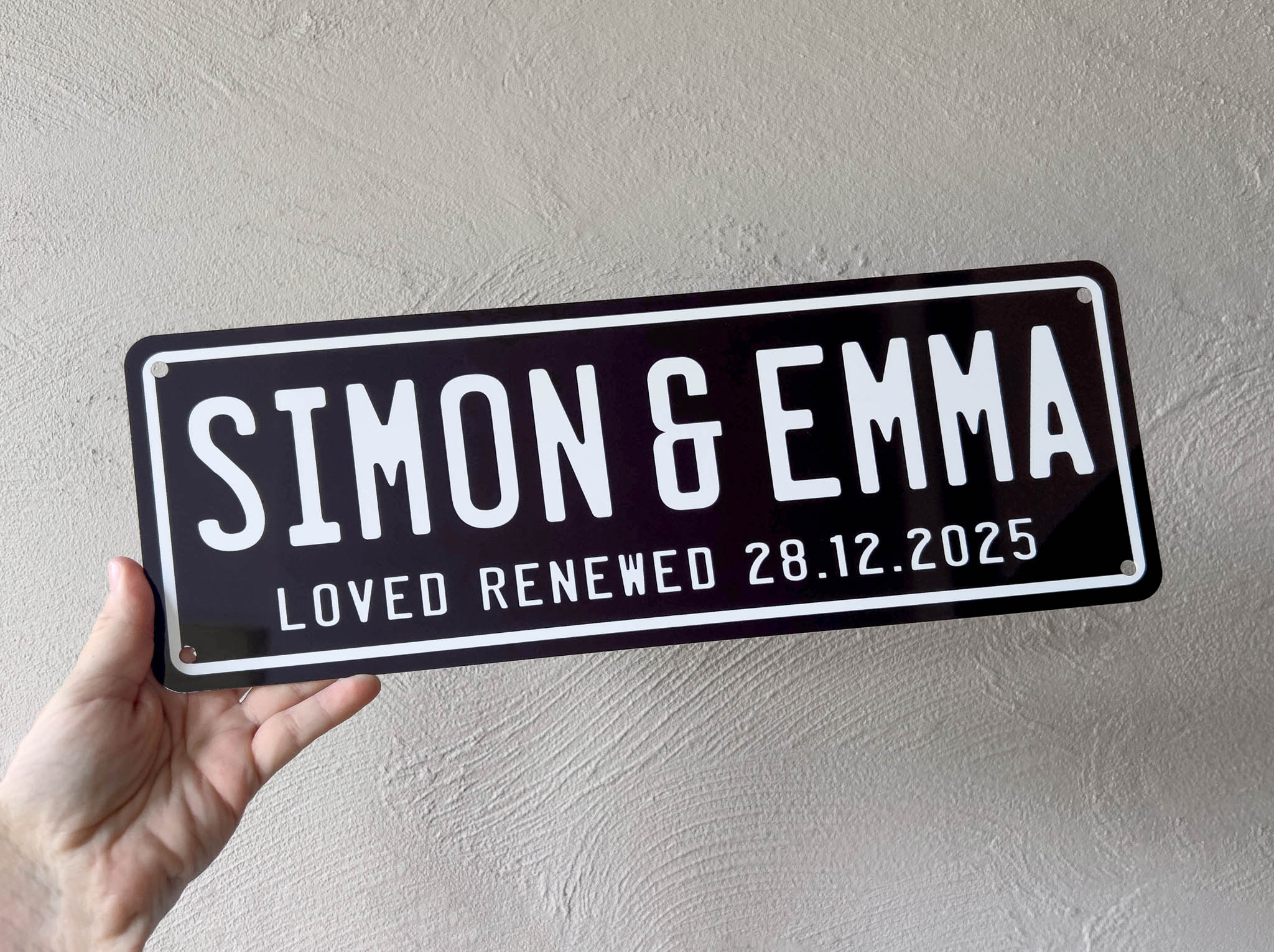 Hand holding a personalized sign with 'Simon & Emma' and a date against a plain background