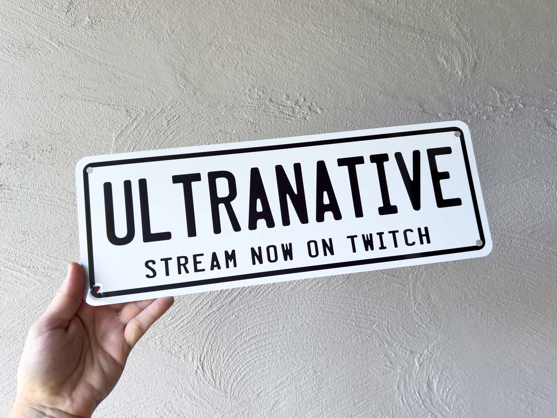 Hand holding a sign with 'ULTRANATIVE' and 'Stream Now on Twitch' text against a plain background