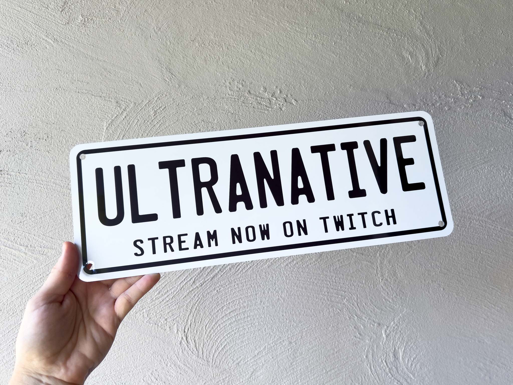 Hand holding a sign with 'ULTRANATIVE' and 'Stream Now on Twitch' text against a plain background