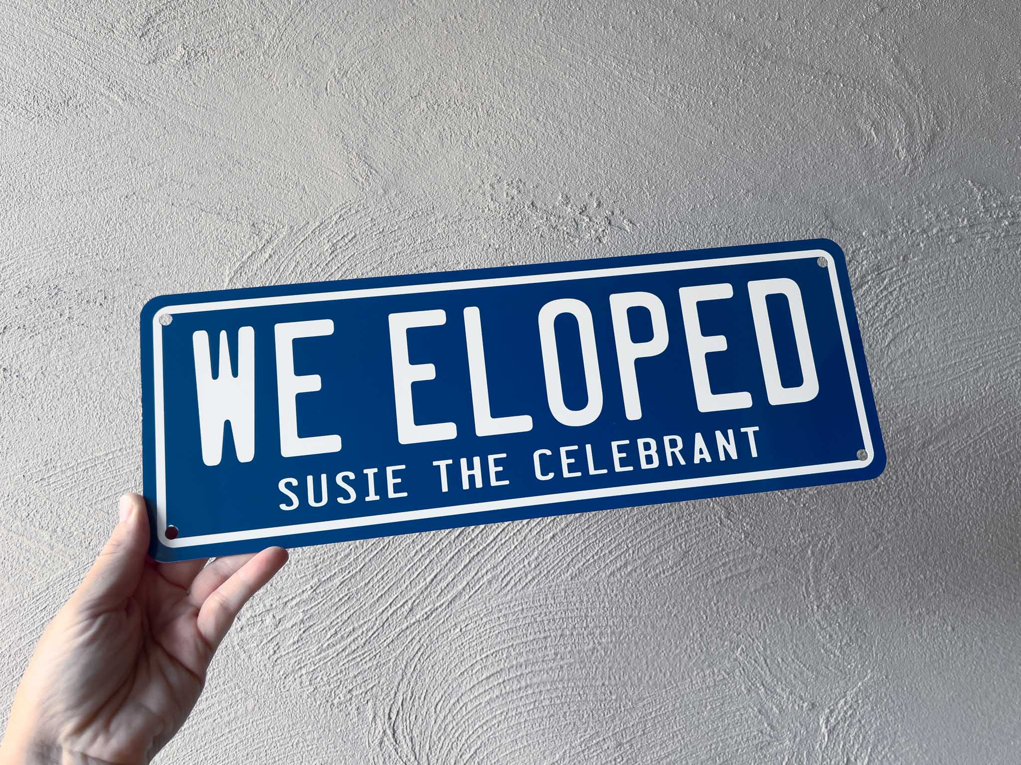 Hand holding a deep blue sign with 'WE ELOPED' and 'Susie the Celebrant' text against a grey background