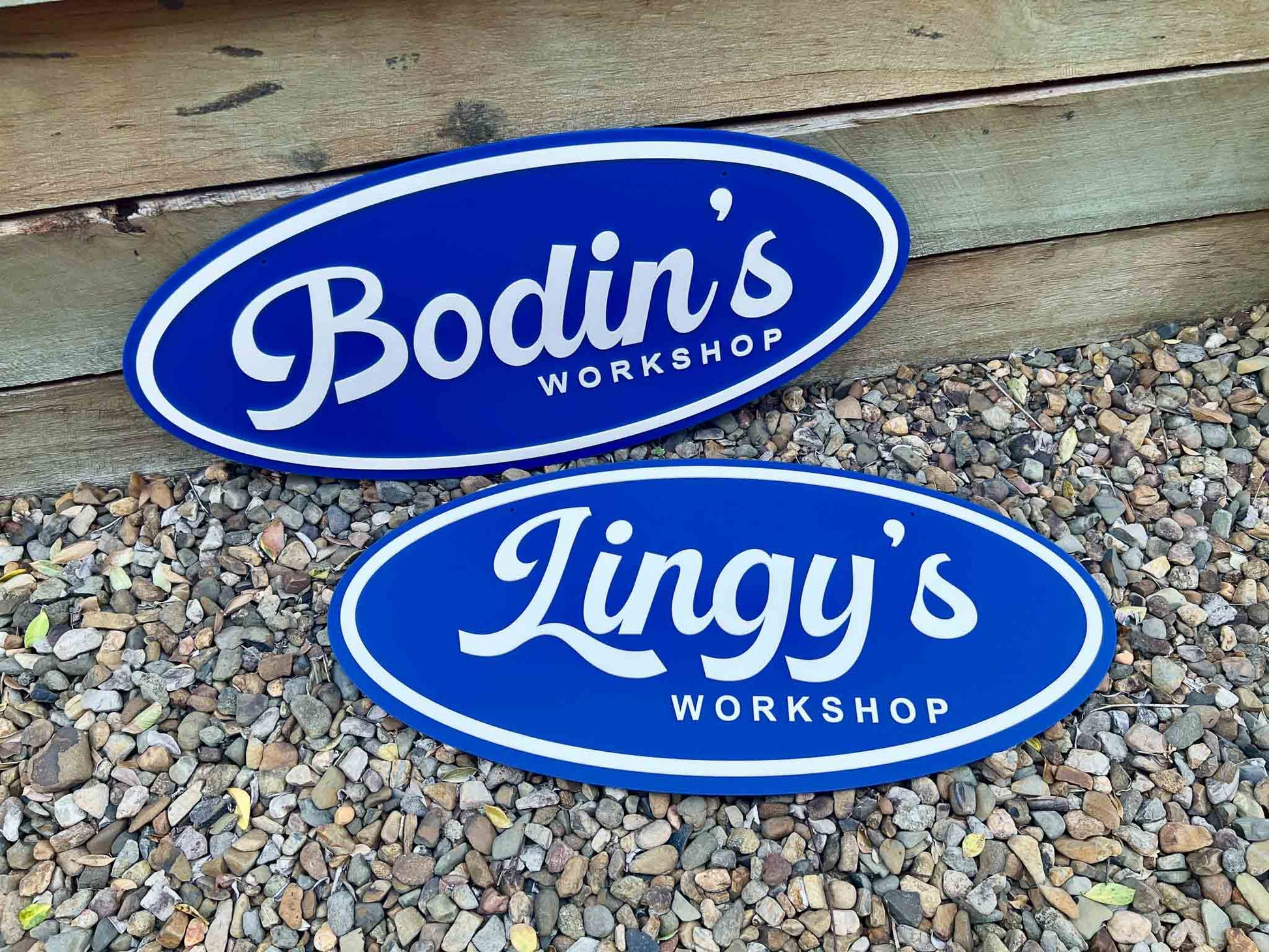 Two oval 3D acrylic signs with 'Bodin's Workshop' and 'Lingy's Workshop' on a wooden and gravel background.