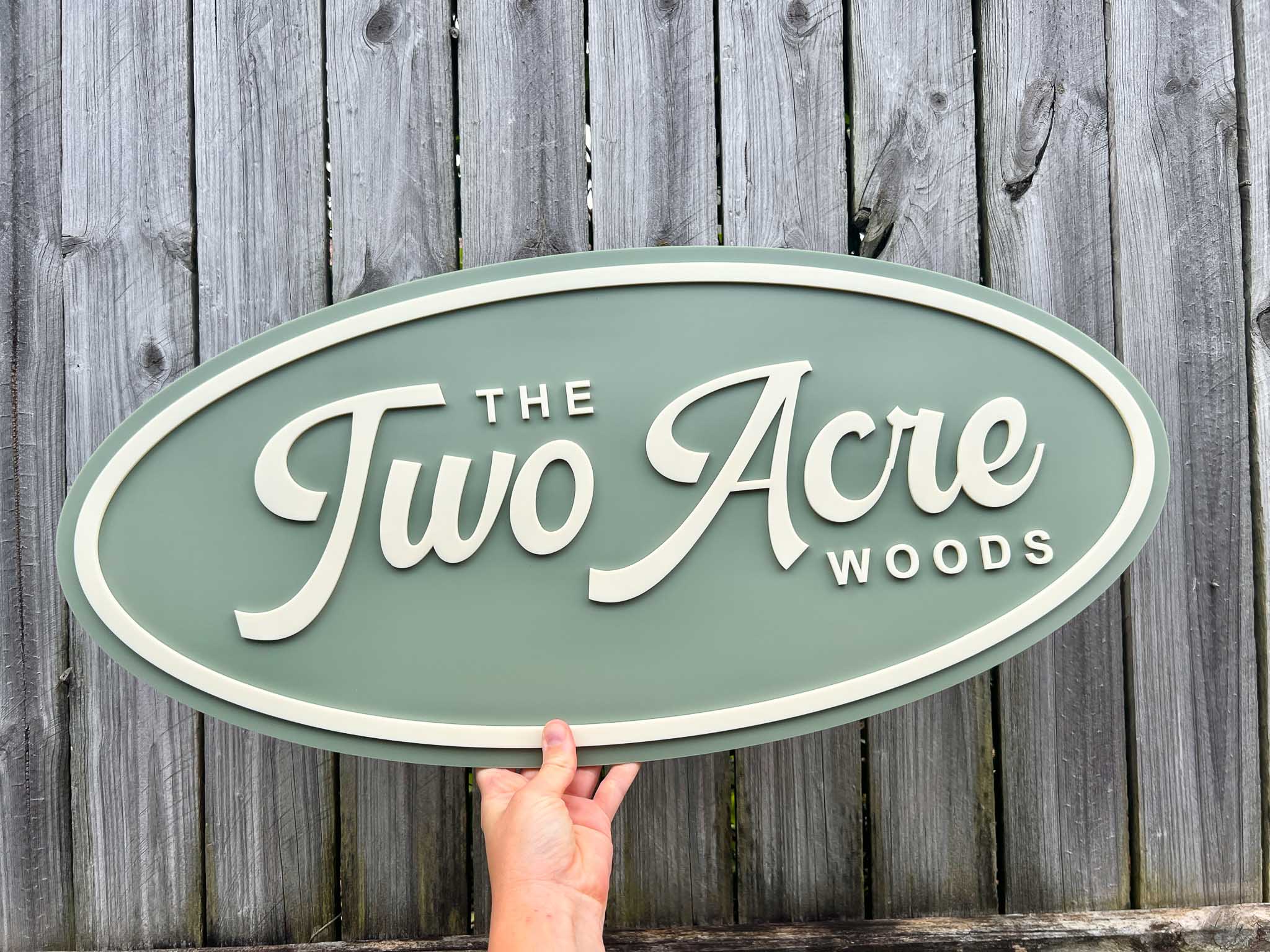 Classic personalised oval sign with 'The Two Acre Woods' text held against a wooden fence background