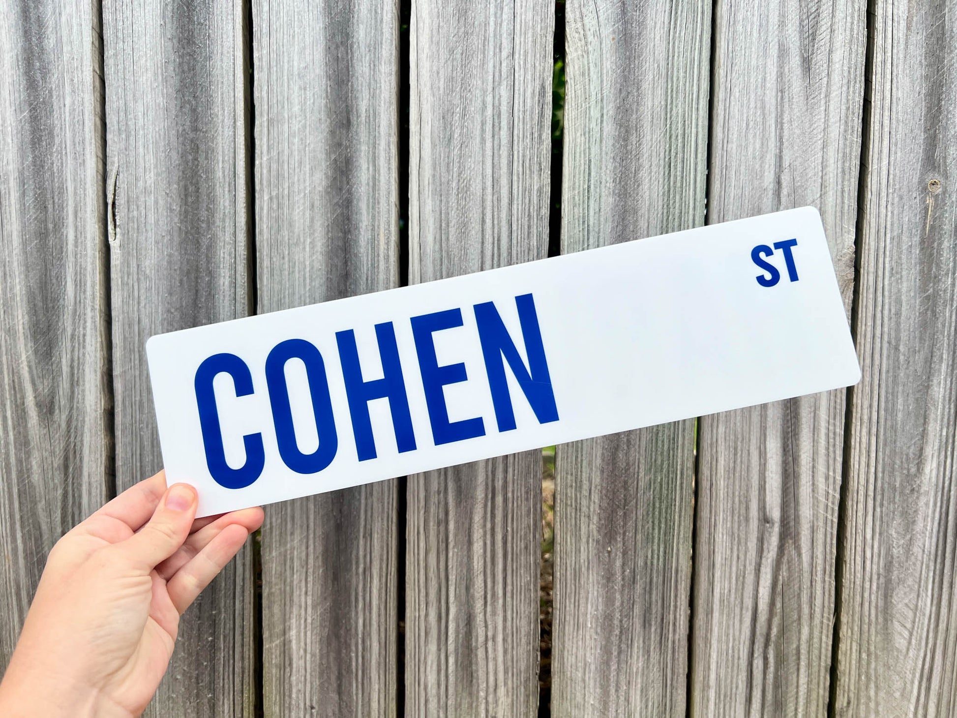 Hand holding a street sign with 'COHEN ST' against a wooden fence background