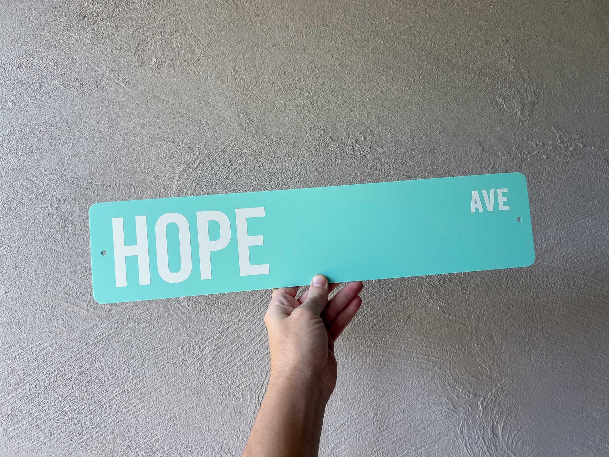 Hand holding a personalised aqua street sign with 'HOPE AVE' on a plain wall.