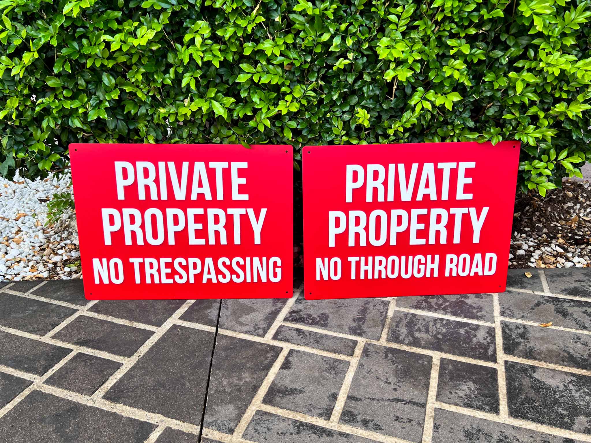 Two red 'Private Property No Trespassing' signs on a stone path with greenery in the background.