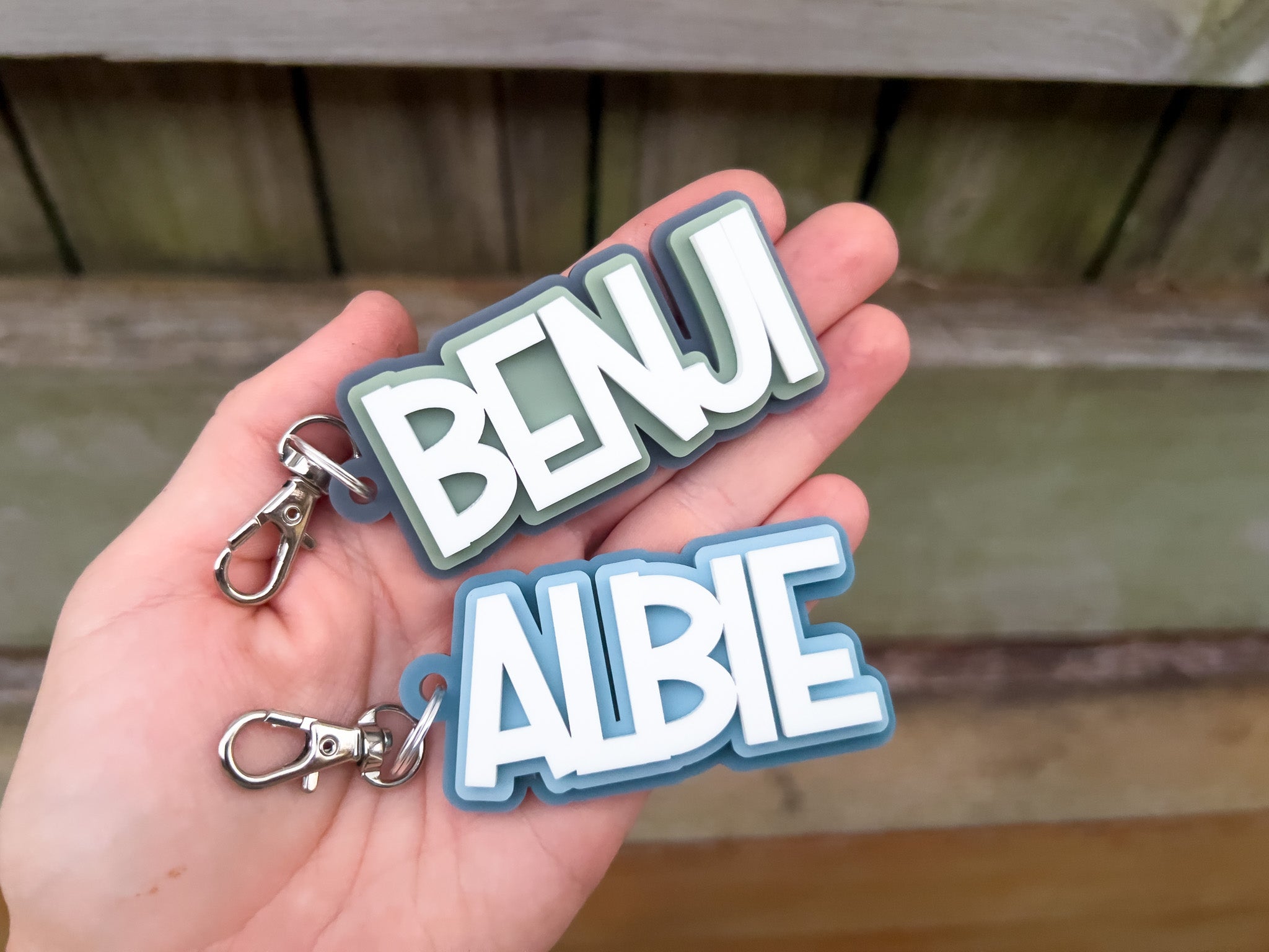 Hand holding two 3D acrylic keychains with 'BENJI' and 'ALBIE' text against a wooden background