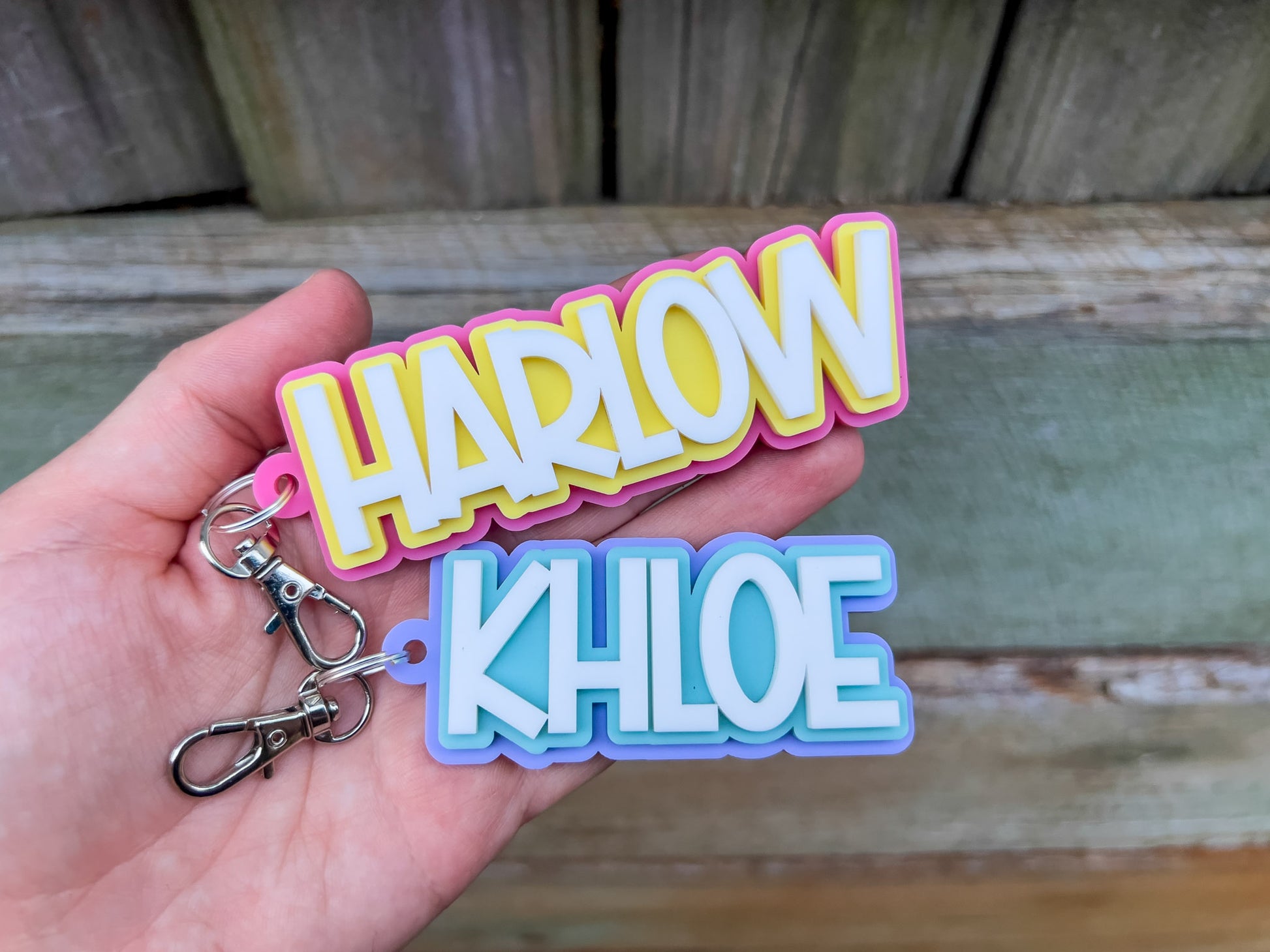 Colorful 3D acrylic 'HARLOW' and 'KHLOE' keychains held in a hand against a wooden background.