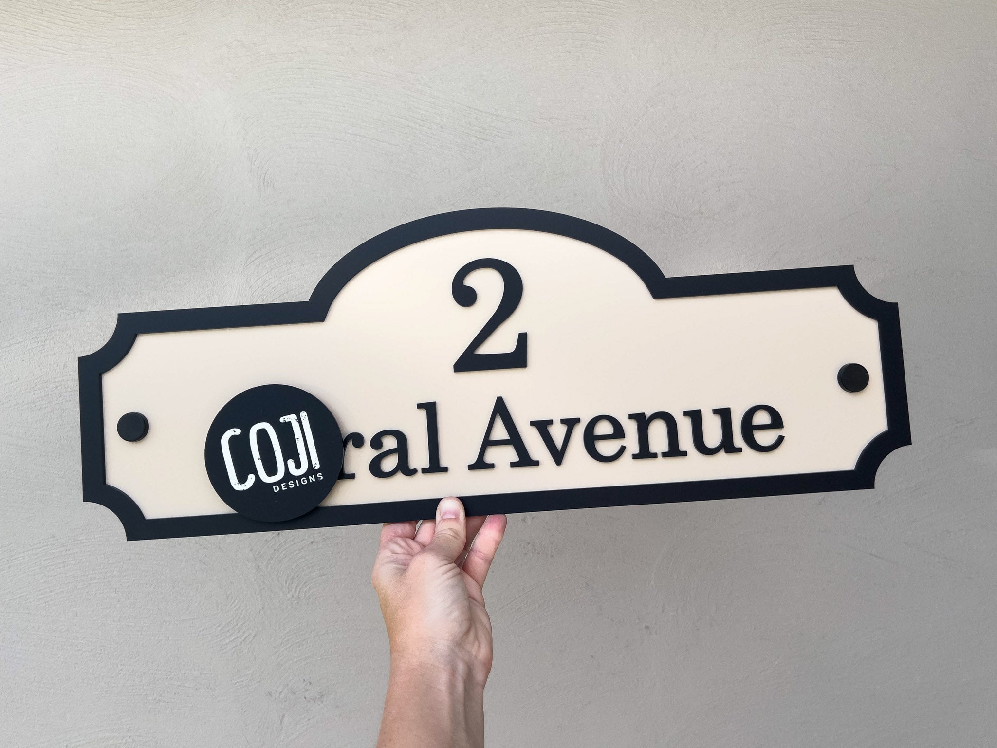 Hand holding a decorative house number sign with '2 Avenue' on a plain background.