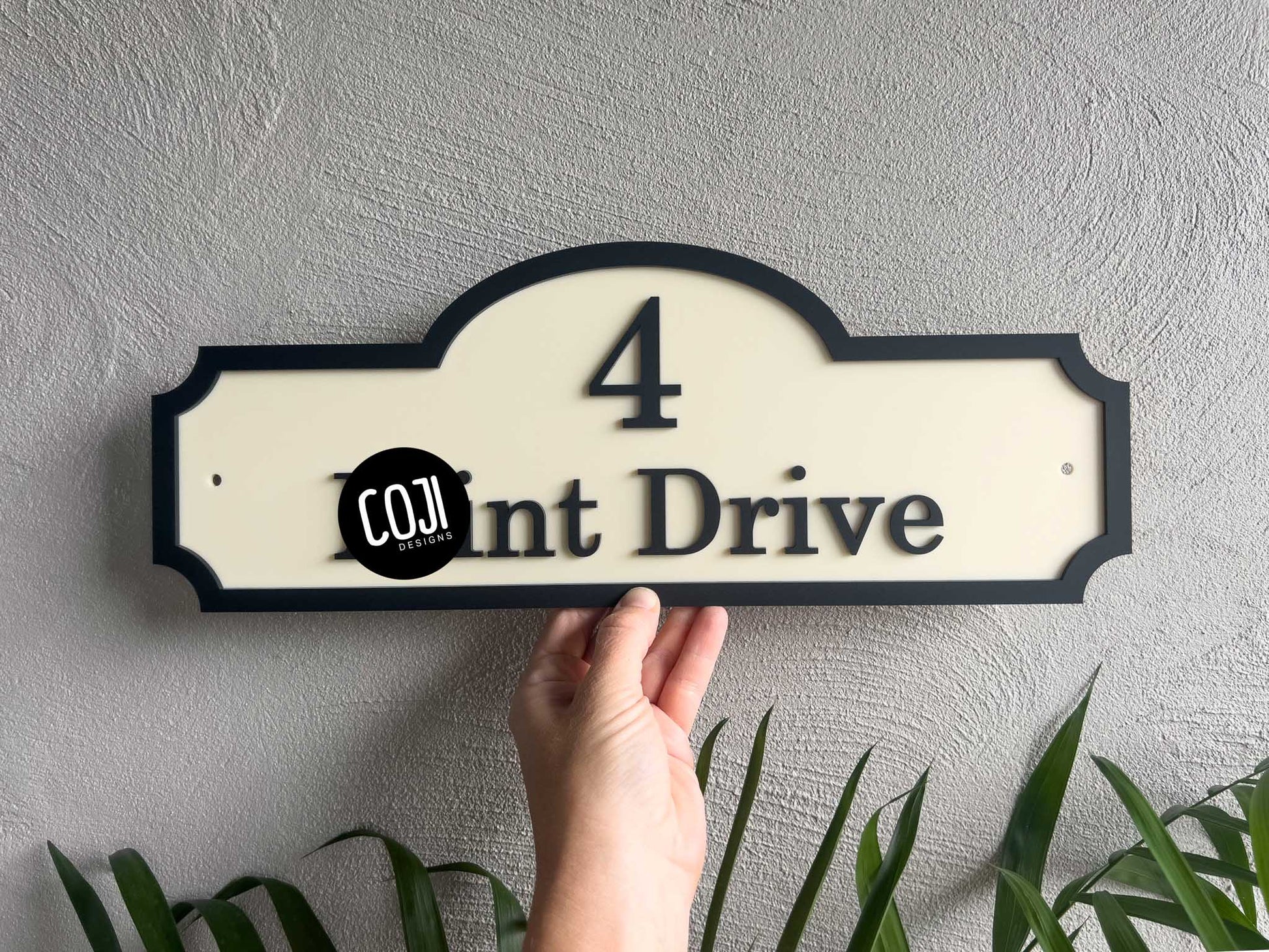 Hand holding a beige and black house number sign with '4 Drive' against a light gray wall.