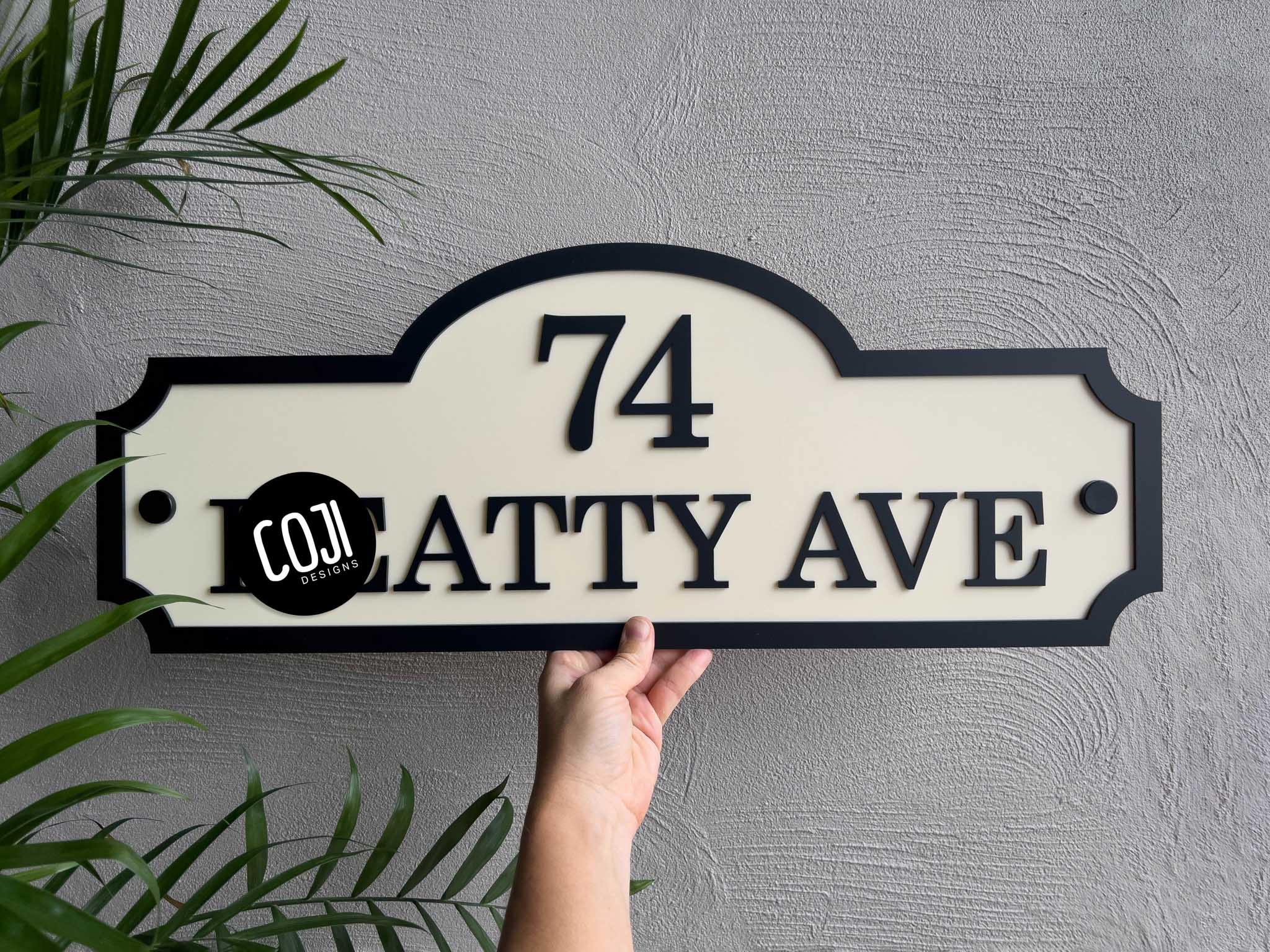 Custom made house number sign '74 Ave' held by a hand against a grey wall with greenery.