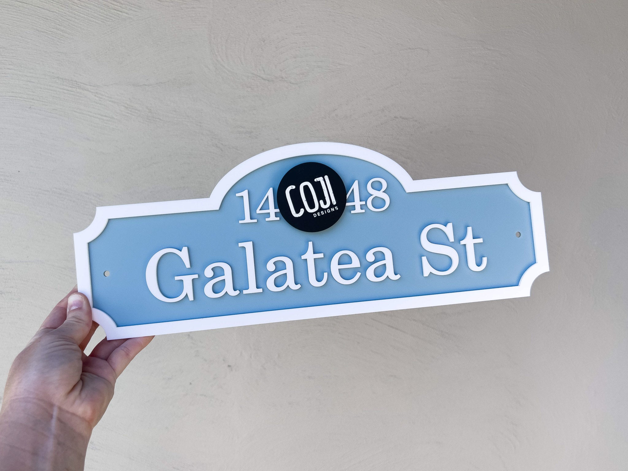 Hand holding a vintage hampton style blue street sign with white text and on a light gray background