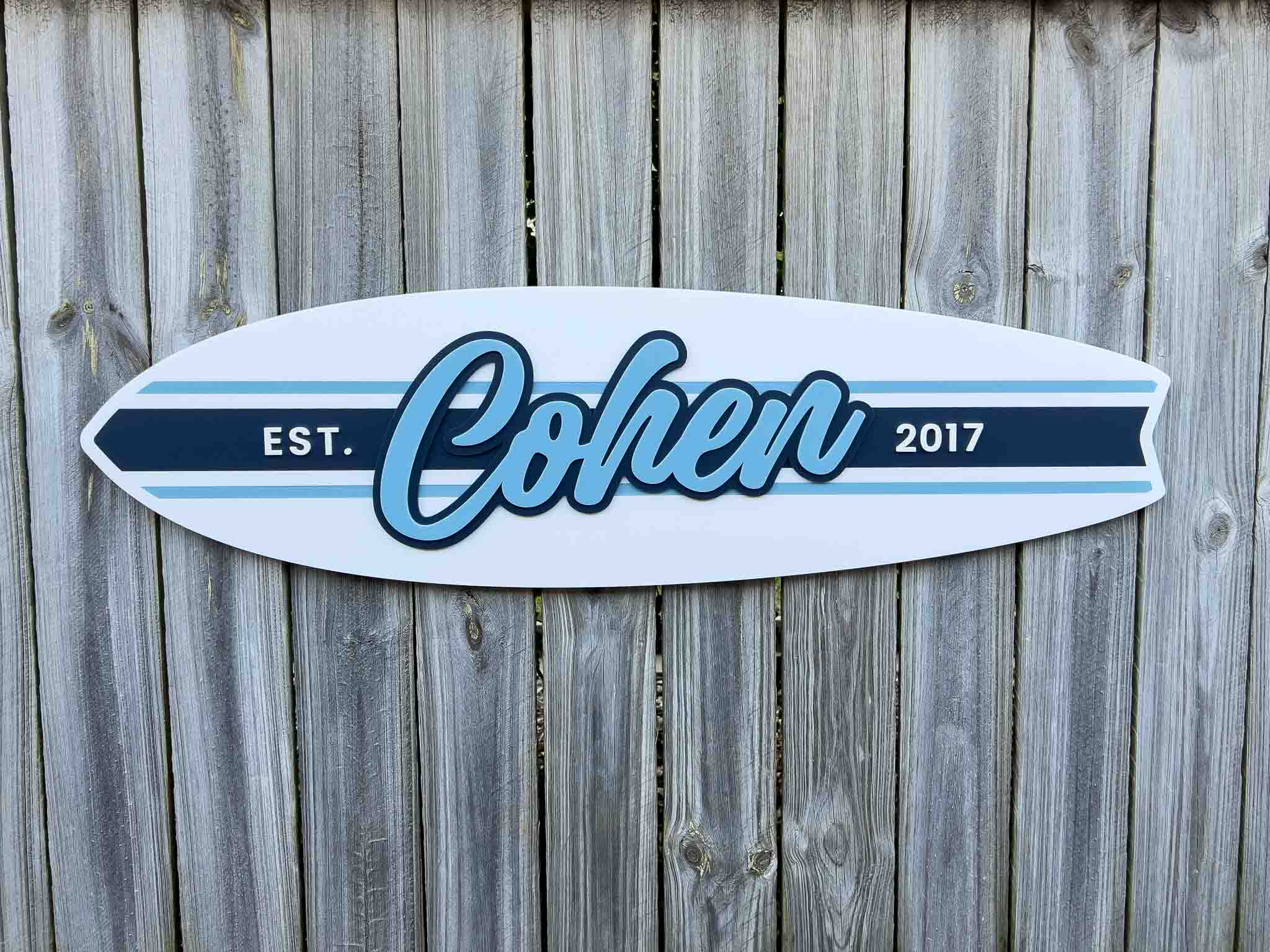 Surfboard-shaped sign with 'Cohen' and 'Est. 2017' on a wooden background