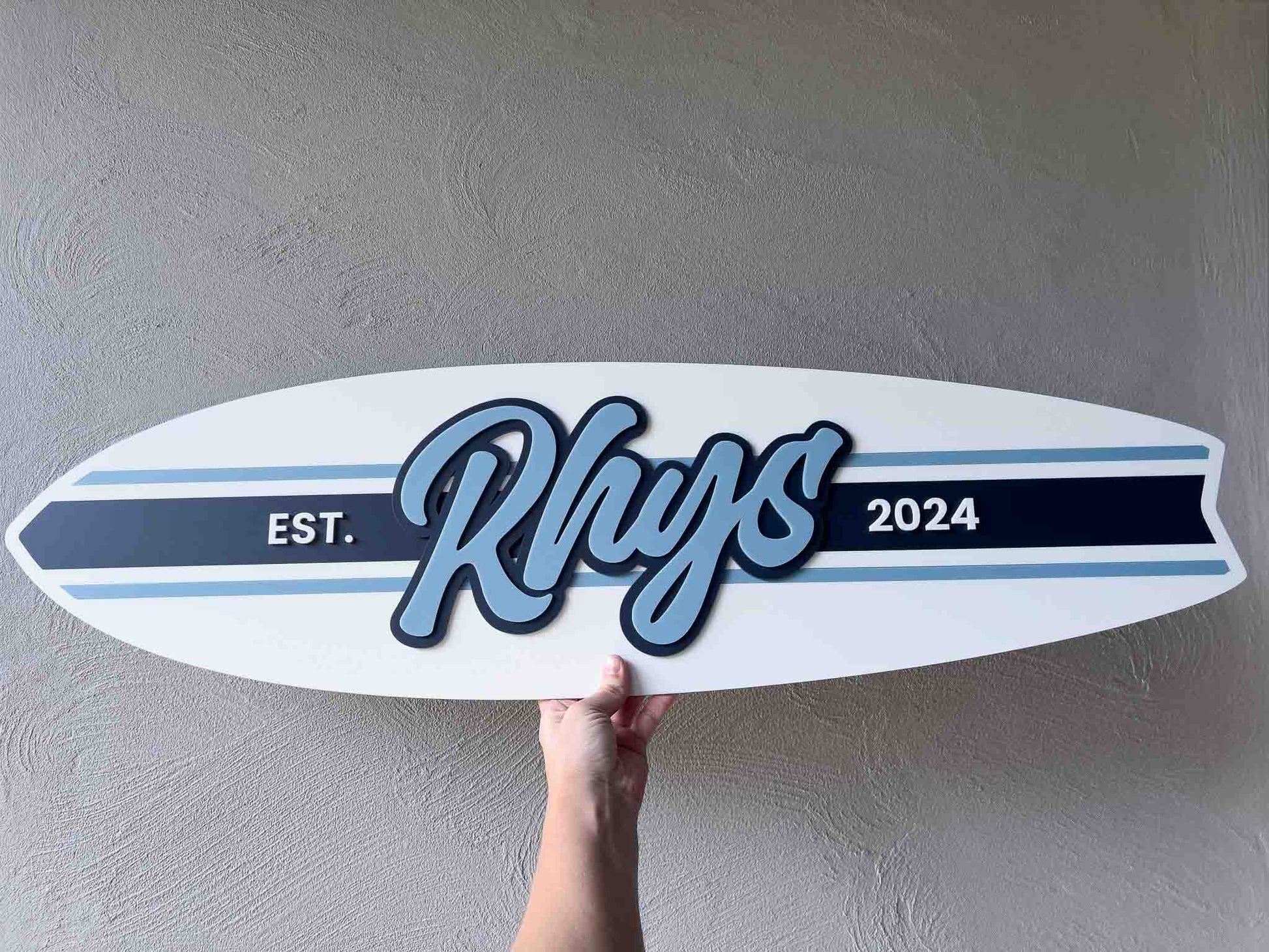 Surfboard-shaped sign with 'Rhys' and '2024' on a plain wall
