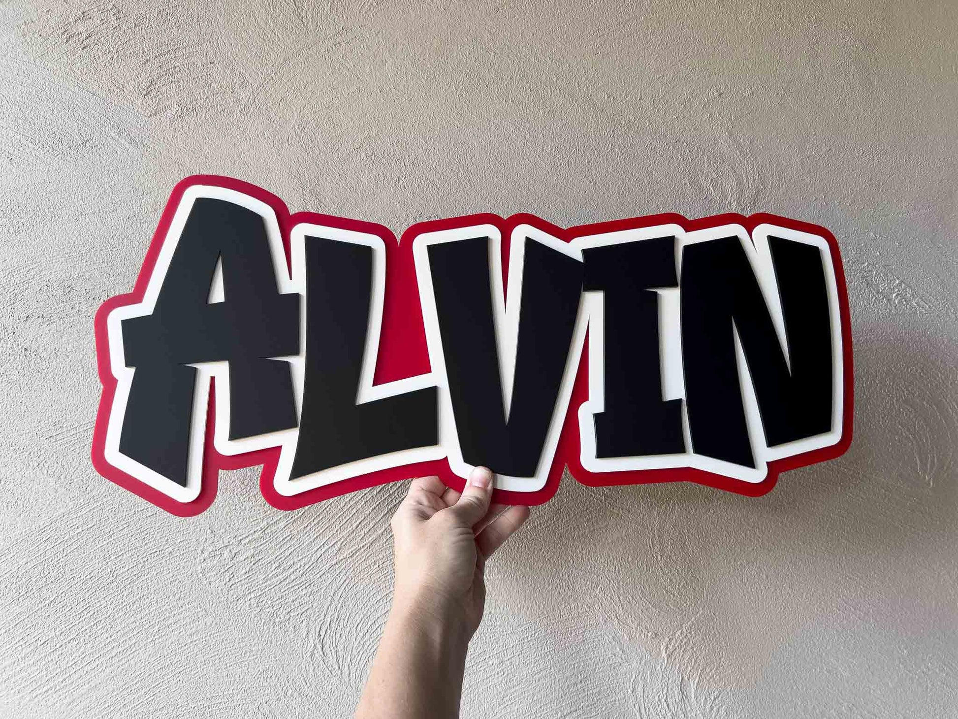 Hand holding a large personalised name plaque with 'ALVIN' written in red, black and white.