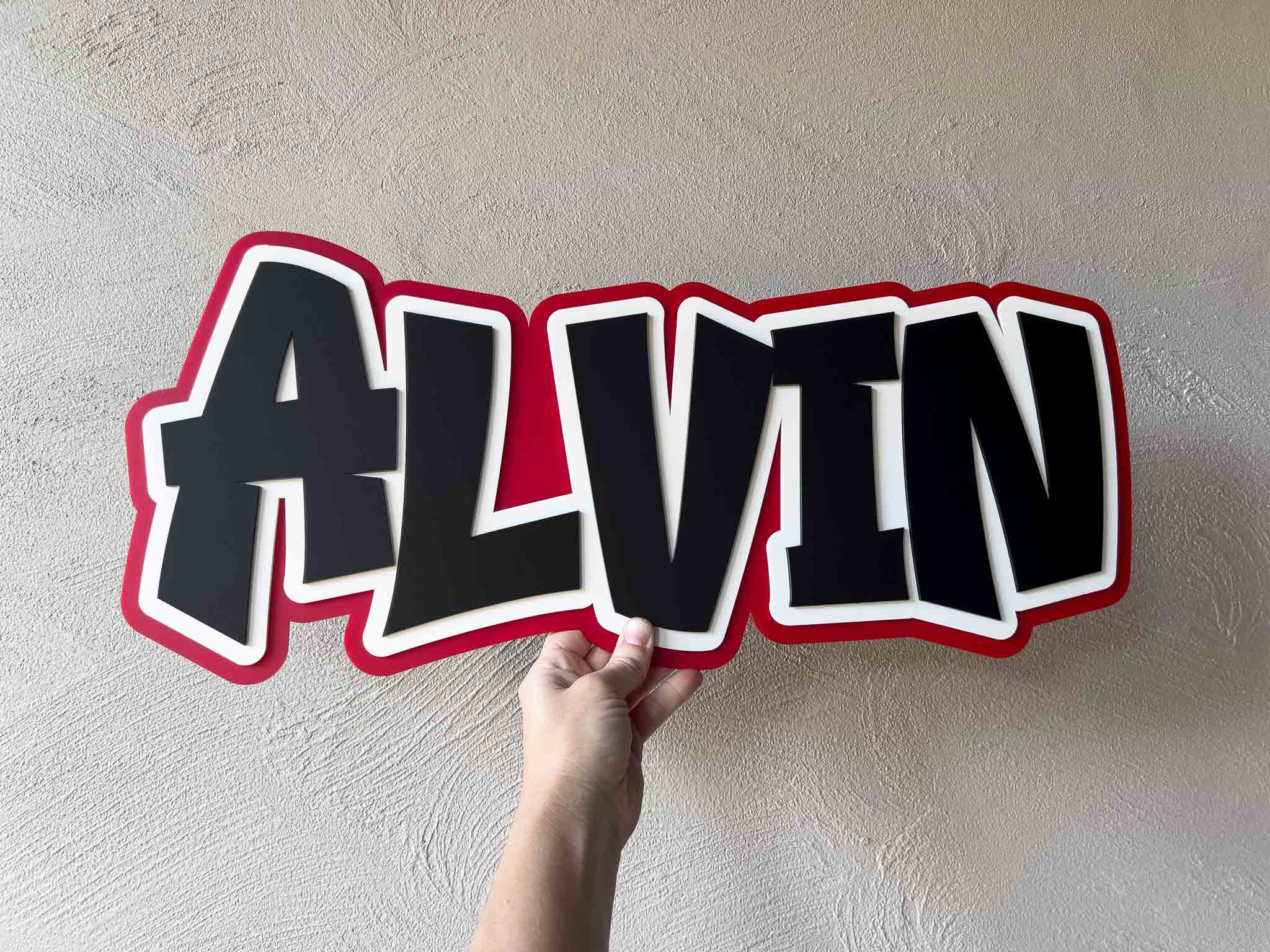 Hand holding a large personalised name plaque with 'ALVIN' written in red, black and white.