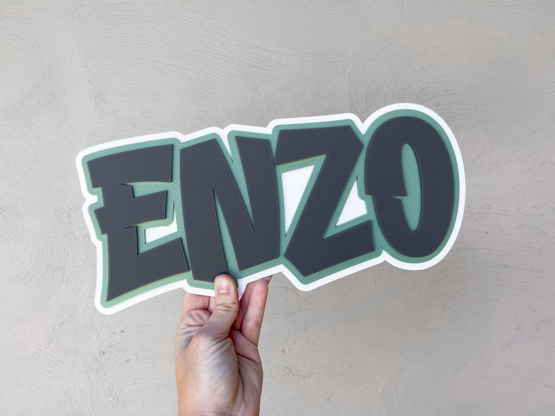 Hand holding a 3D name sign with 'ENZO' in white, green and grey.