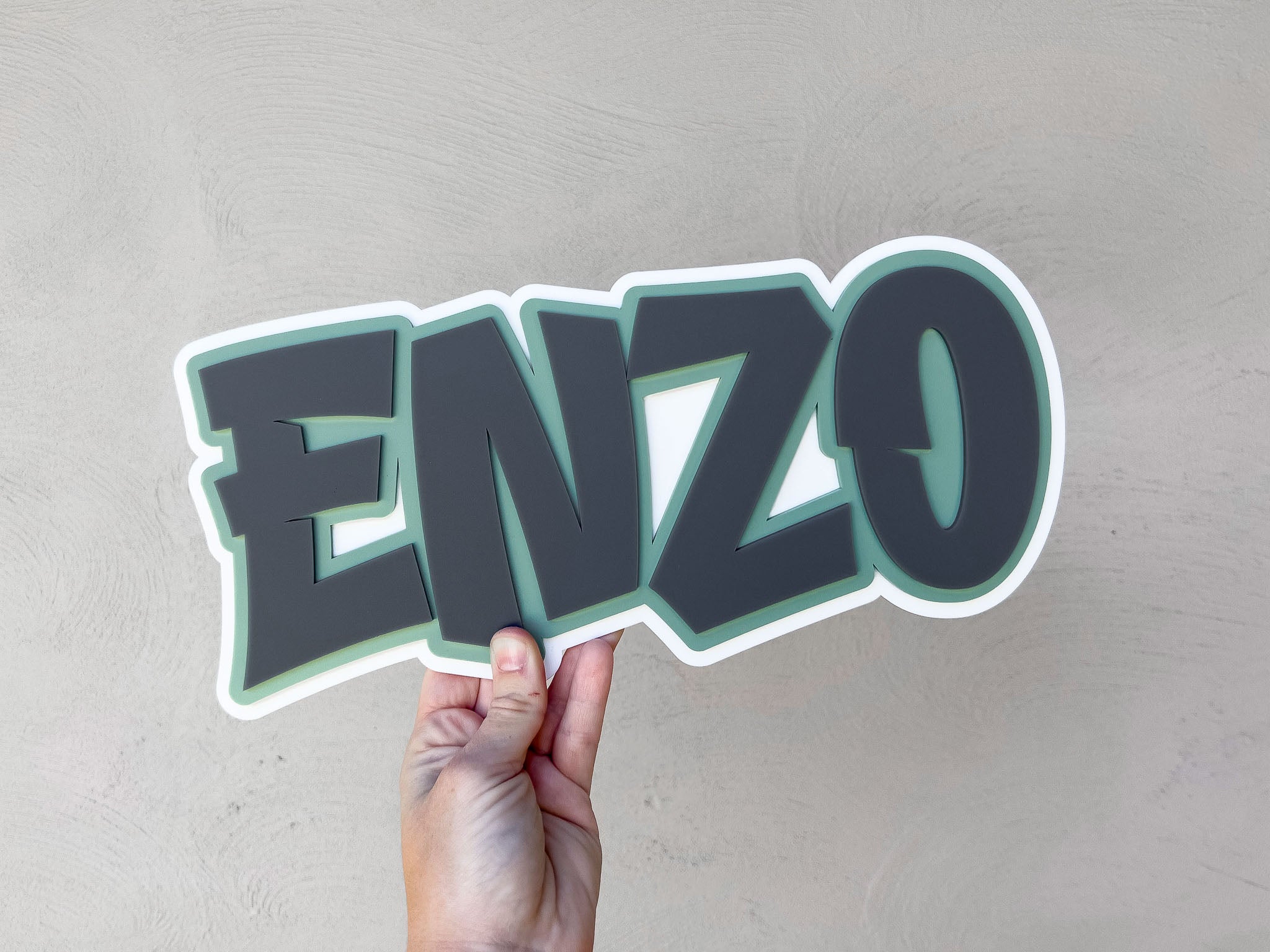 Hand holding a 3D name sign with 'ENZO' in white, green and grey.