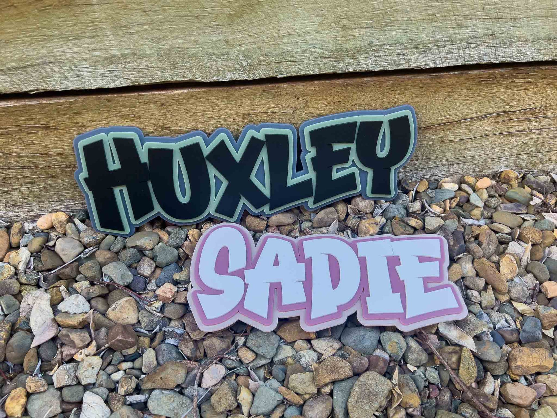 Two acrylic name plaques, 'HUXLEY' and 'SADIE', displayed on a pebble bed with a wooden background.