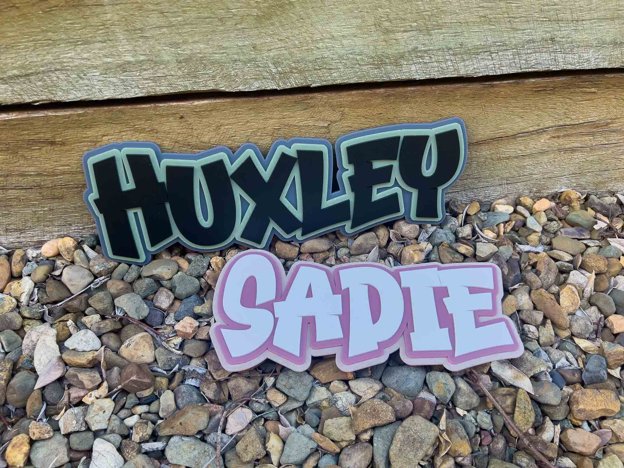 Two acrylic name plaques, 'HUXLEY' and 'SADIE', displayed on a pebble bed with a wooden background.