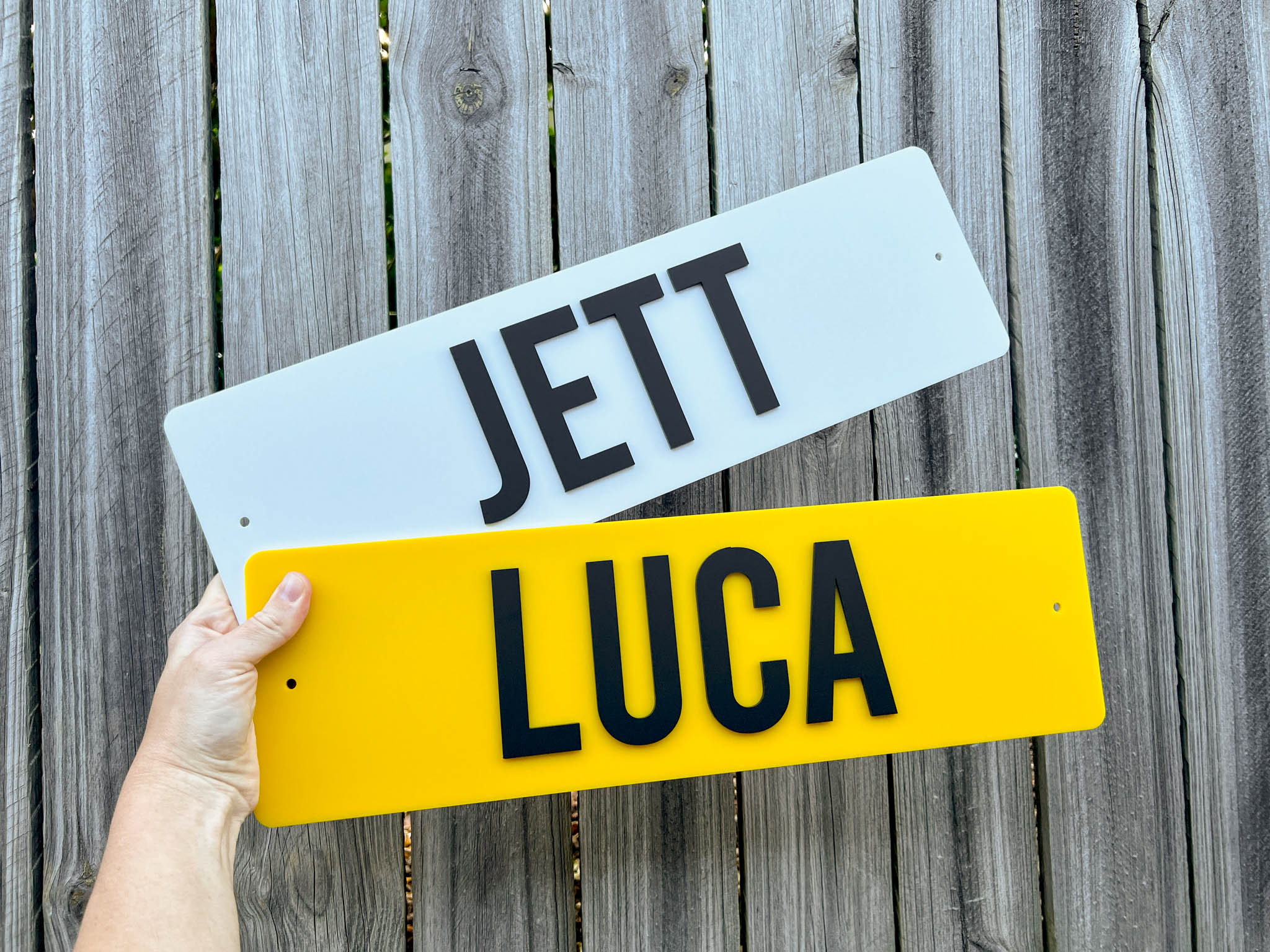 Two surf style name plate-style signs, one white with 'Jett' and one yellow with 'Luca', held against a wooden fence.