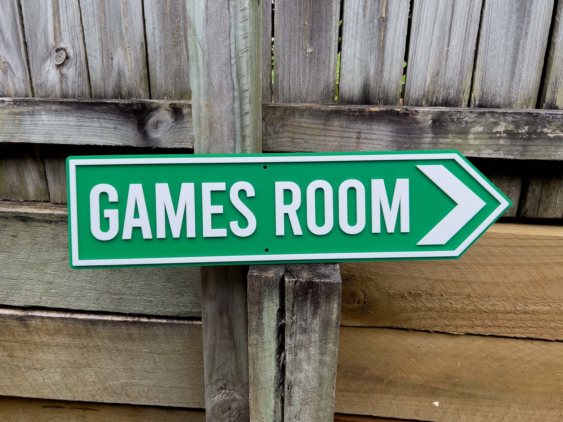 Green 'GAMES ROOM' sign with white text and arrow on wooden post against a wooden fence background | Coji Designs