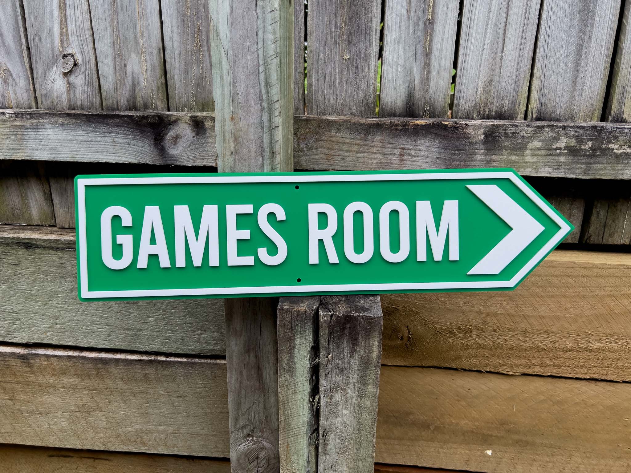Green 'GAMES ROOM' sign with white text and arrow on wooden post against a wooden fence background | Coji Designs