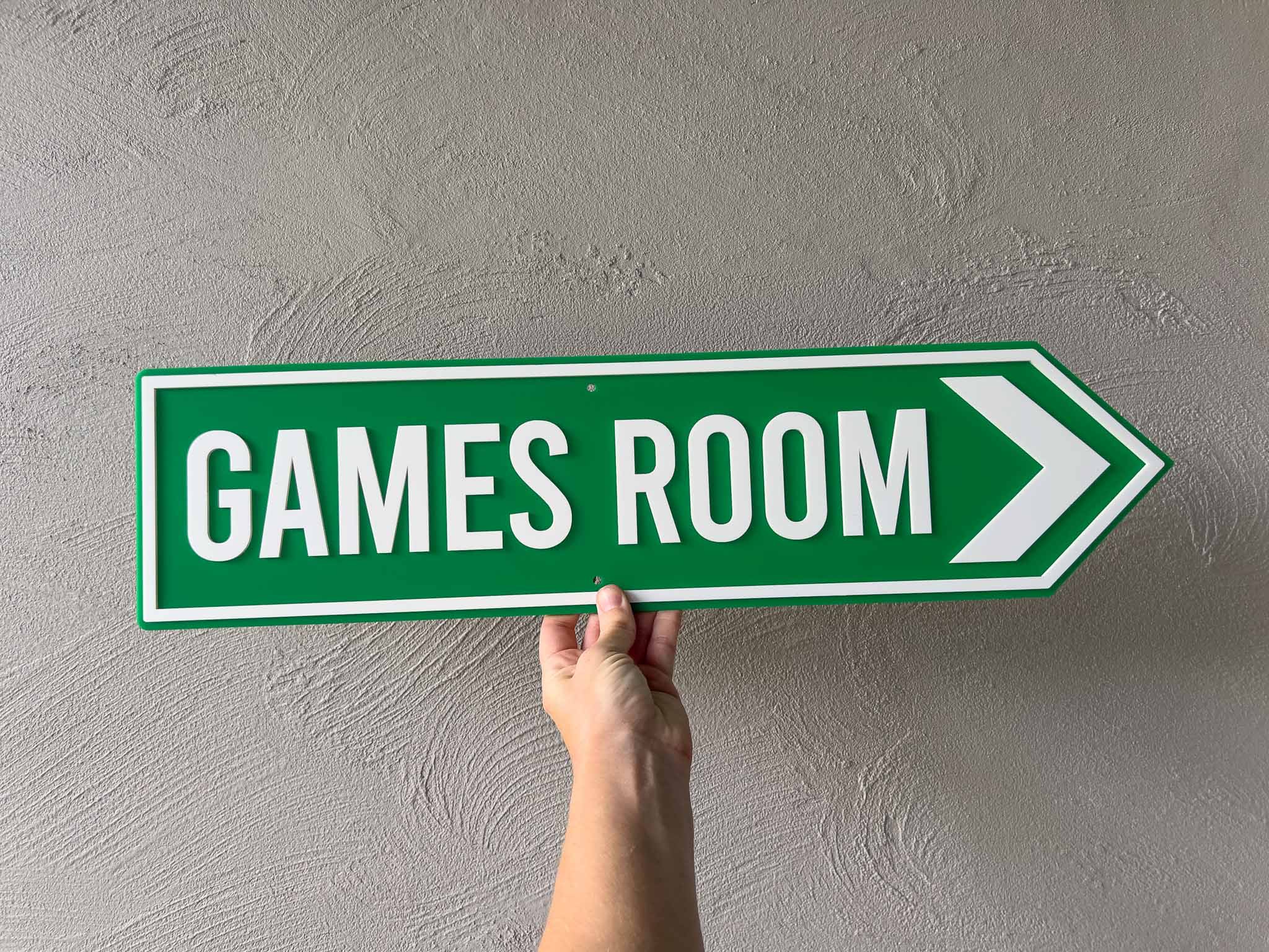 Hand holding a green sign with 'GAMES ROOM' text against a gray wall | Coji Designs