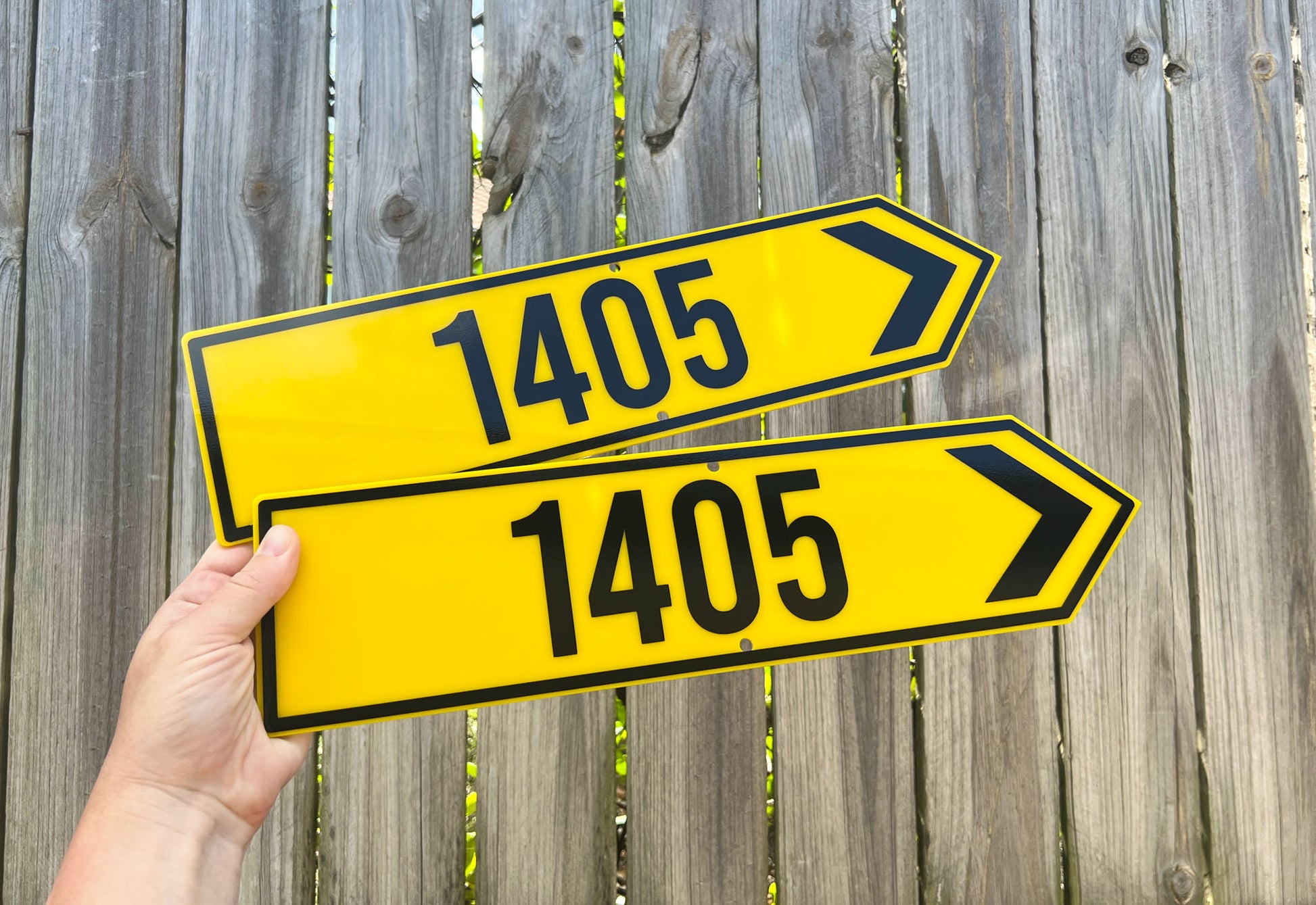 Two yellow directional signs with black text and arrows that read '1405' are held against a wooden fence.