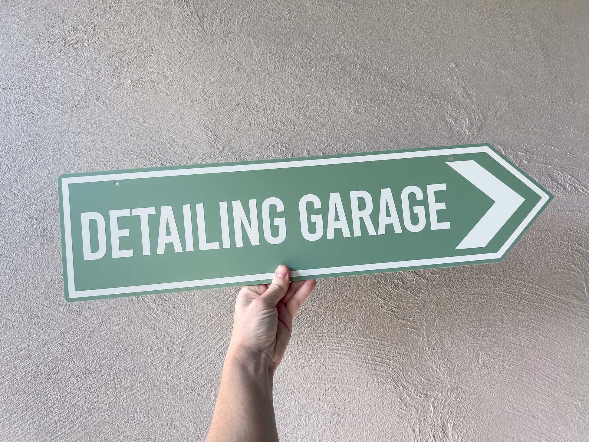 Hand holding a green street sign with 'Detailing Garage' text against a plain wall.