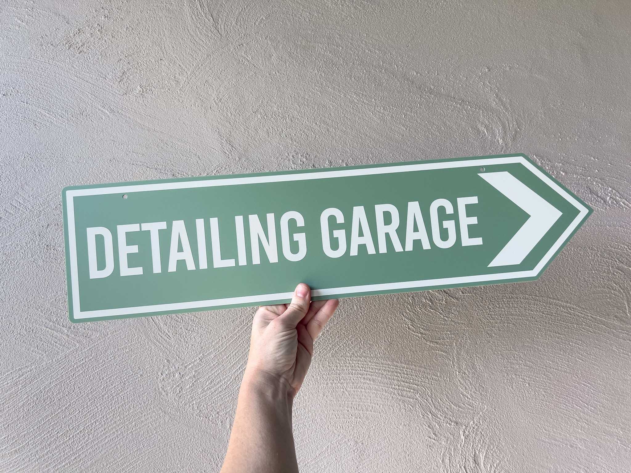 Hand holding a green street sign with 'Detailing Garage' text against a plain wall.
