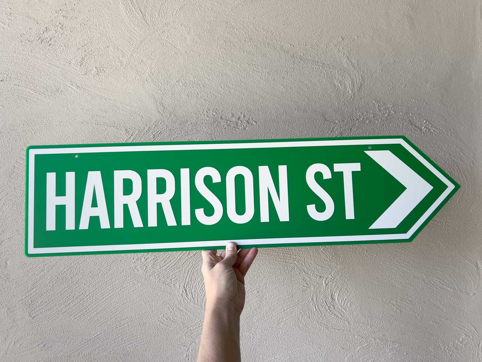Hand holding a vintage green street sign with 'HARRISON ST' text against a plain background