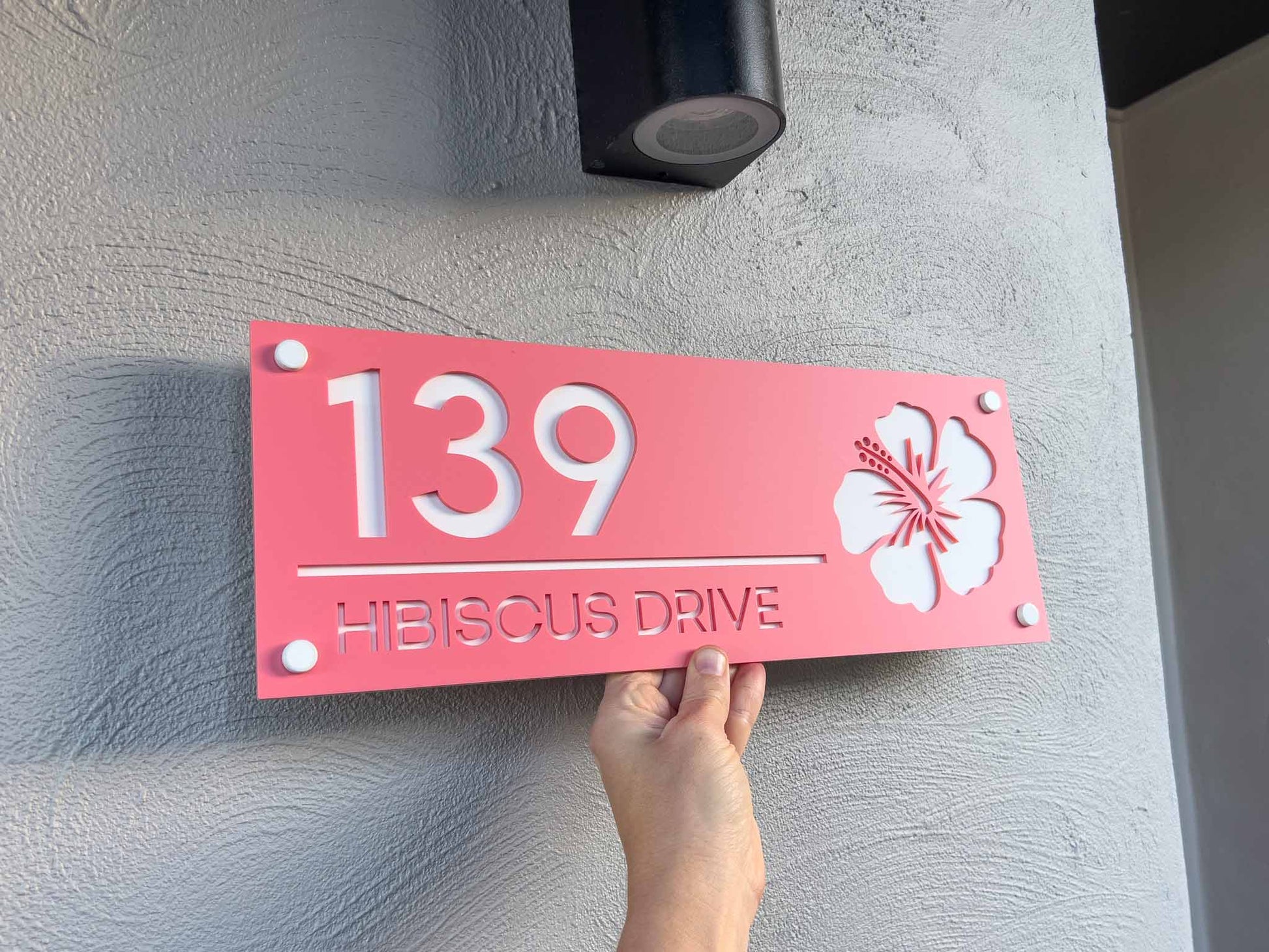 Pink hibiscus house number sign with '139' and 'HIBISCUS DRIVE' text, held by a hand against a light gray wall.
