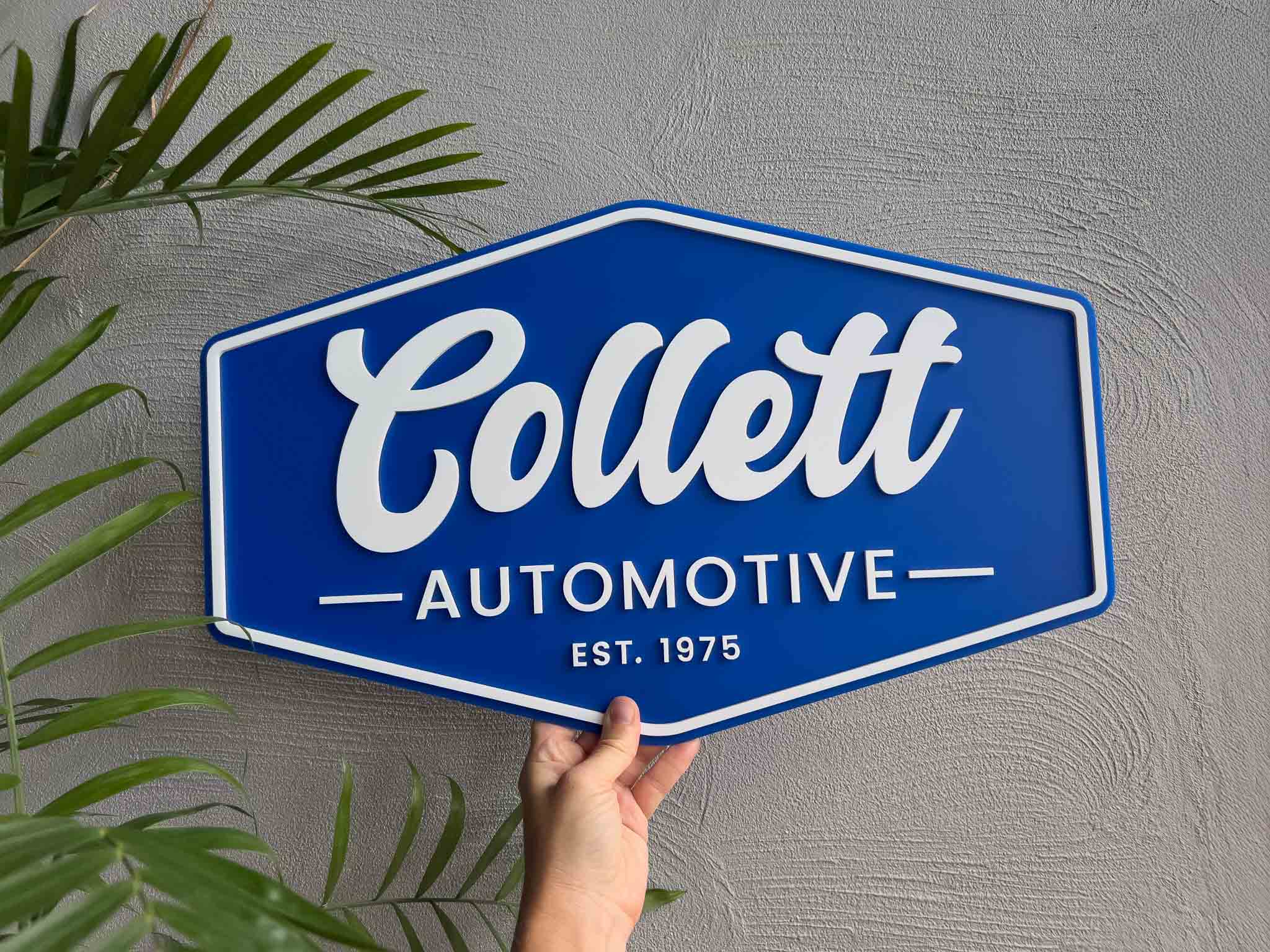 Blue and white custom made sign for 'Collett Automotive' sign held against a gray wall with green leaves.