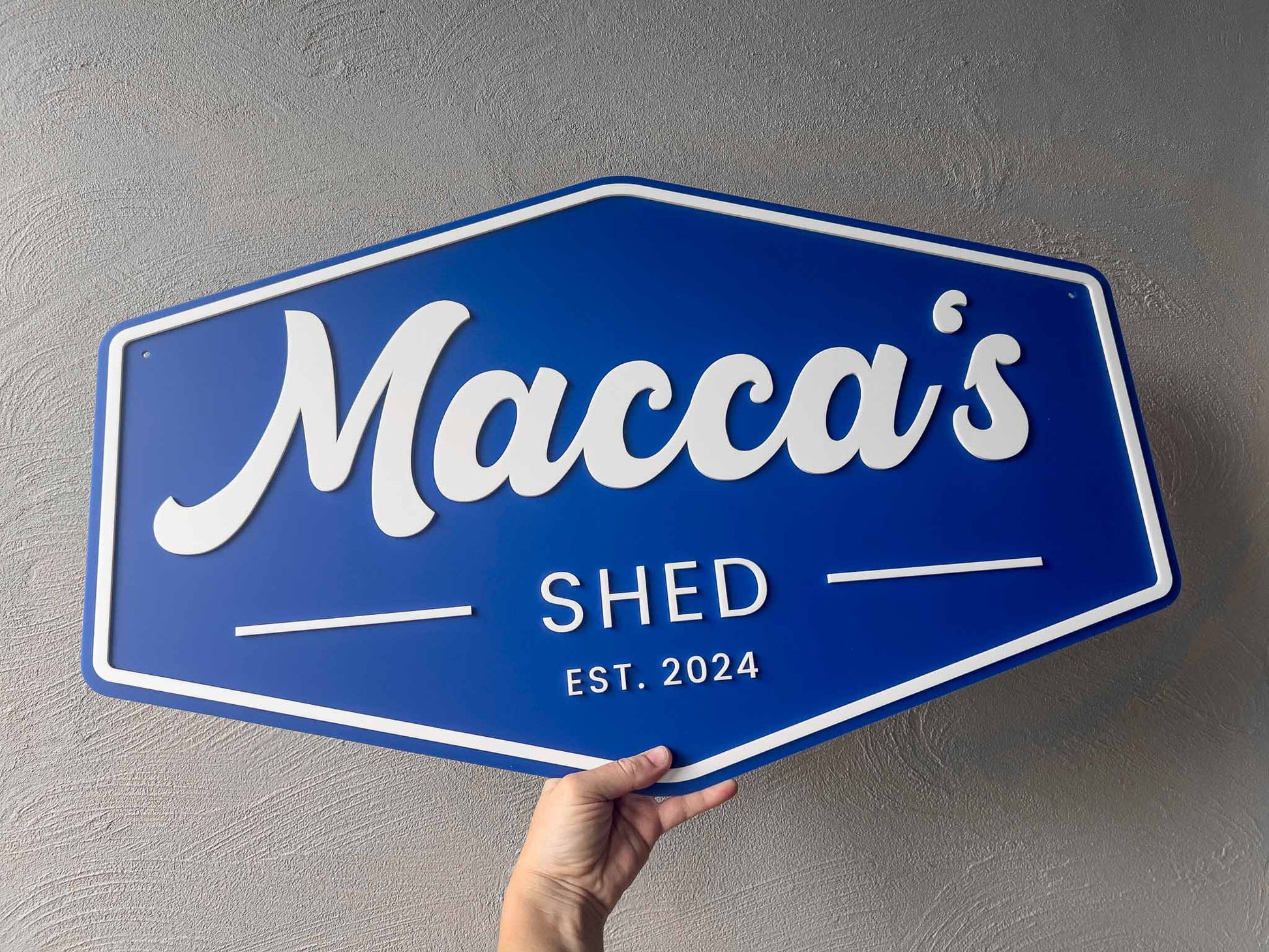 Blue and white custom made sign with 'Macca's Shed Est. 2024' held against a grey wall