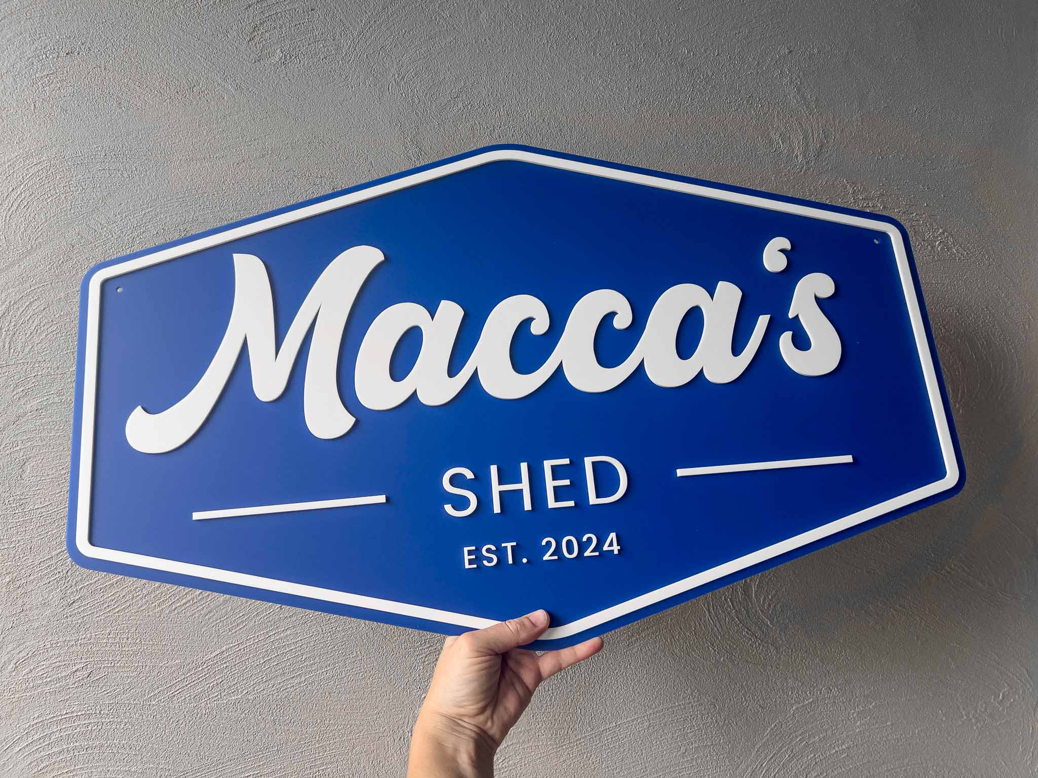 Blue and white custom made sign with 'Macca's Shed Est. 2024' held against a grey wall