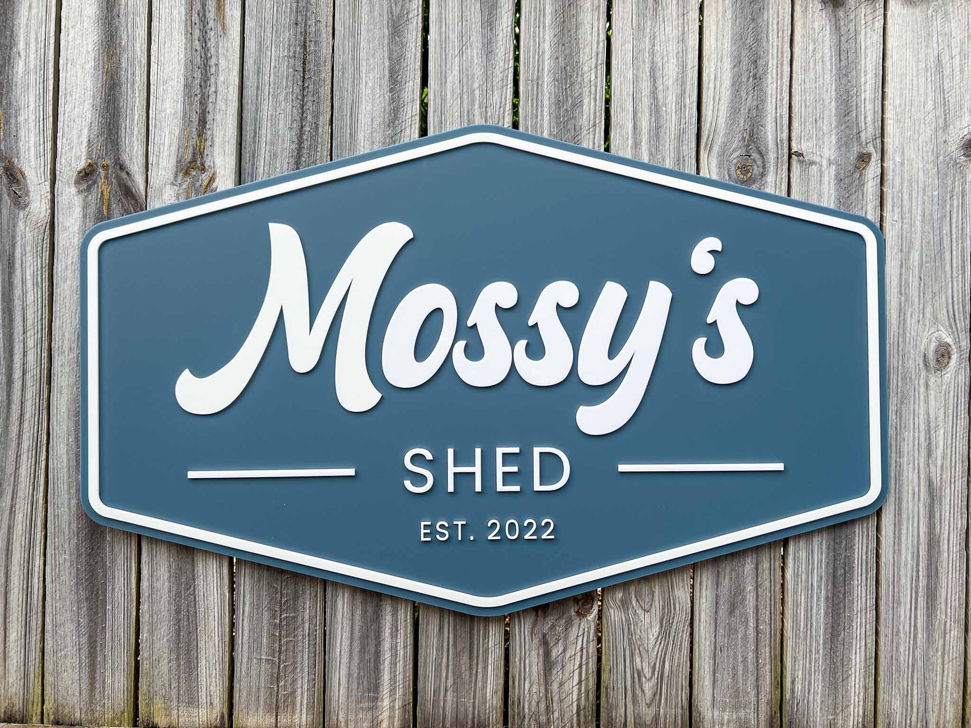 Retro badge style sign with a dusty sea blue background and white text that says 'Mossy's Shed EST. 2022'.