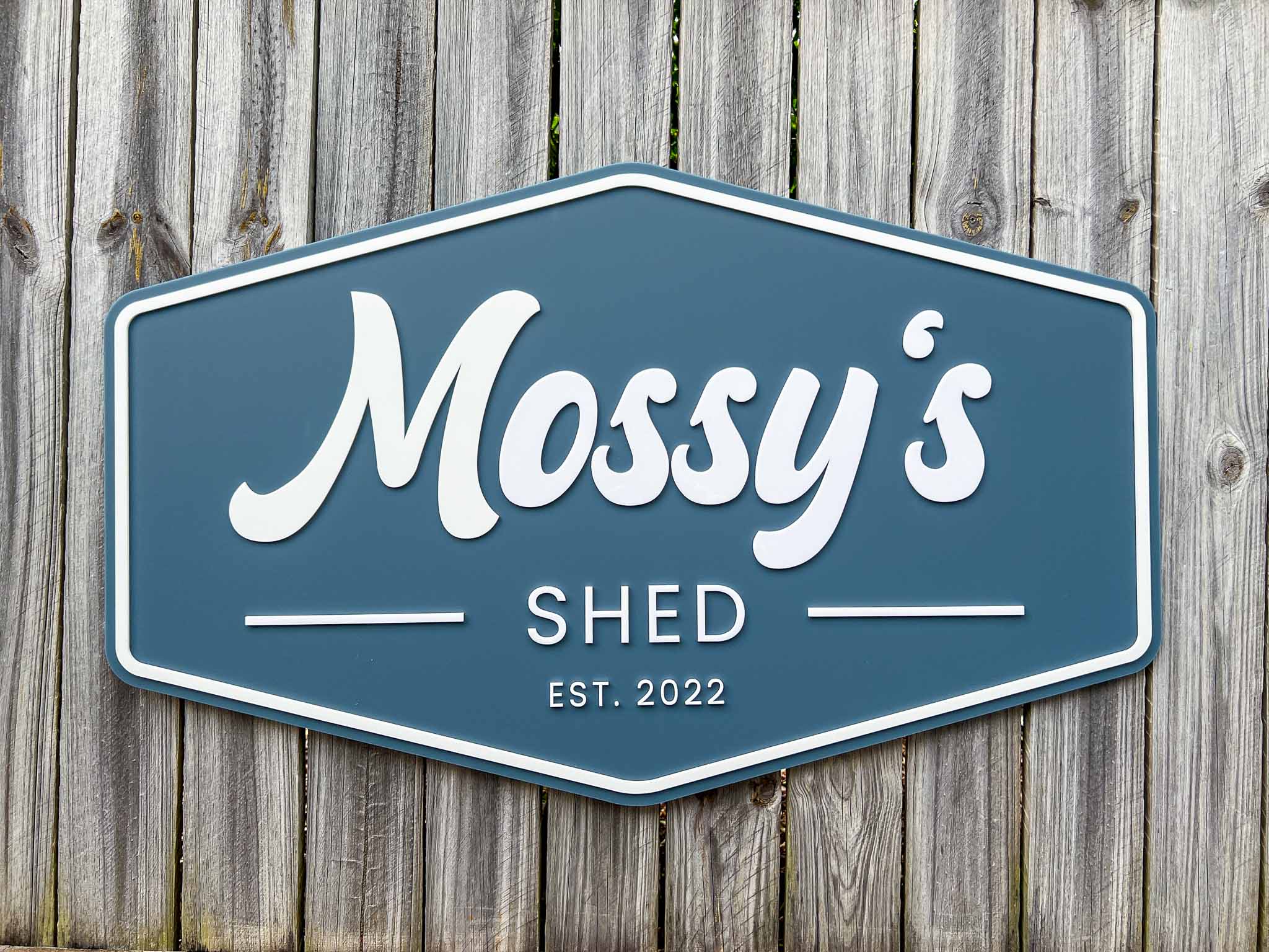 Retro badge style sign with a dusty sea blue background and white text that says 'Mossy's Shed EST. 2022'.
