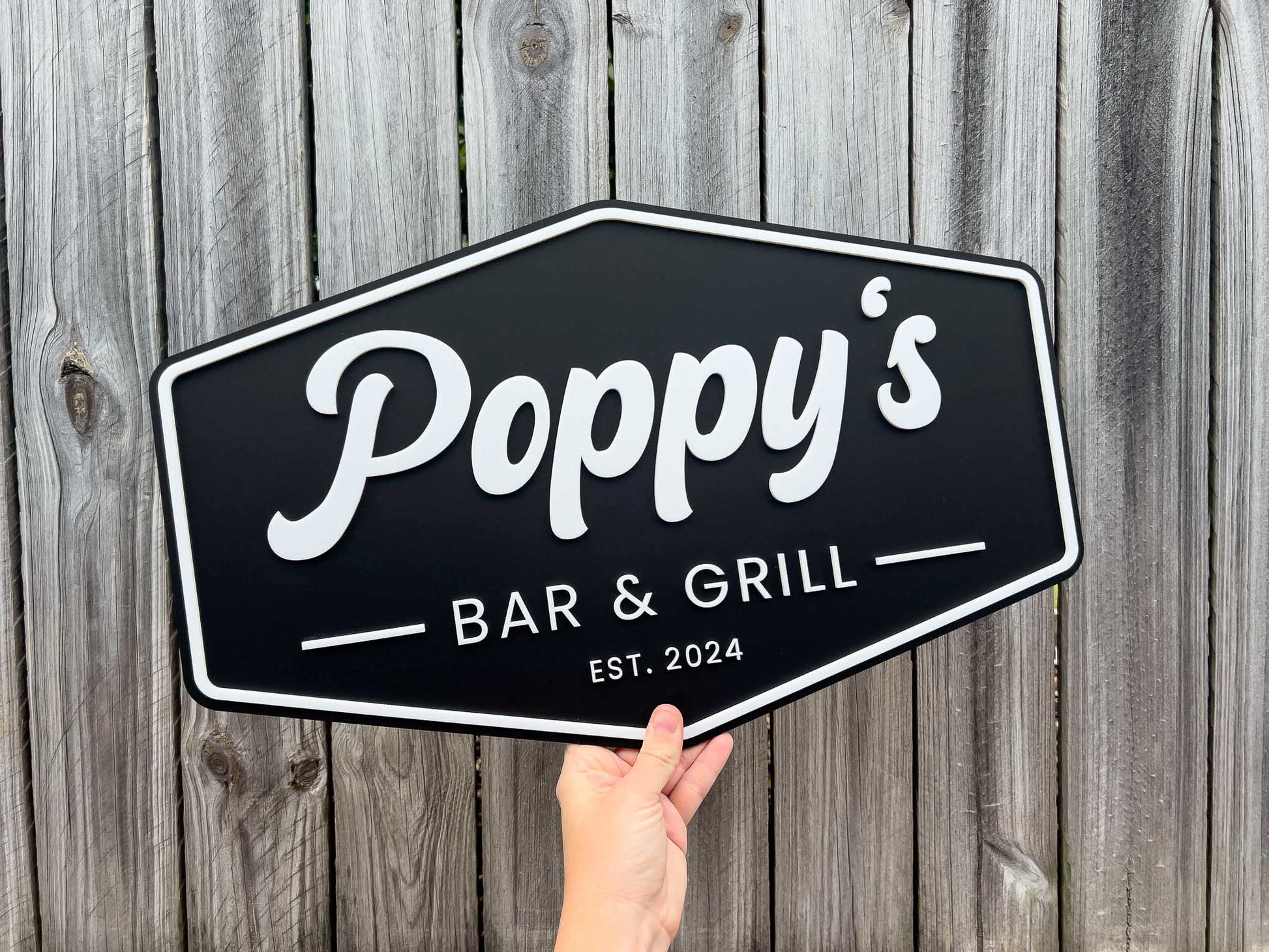 Hand holding a badge style signboard sign with 'Poppy's Bar & Grill' text against a wooden background