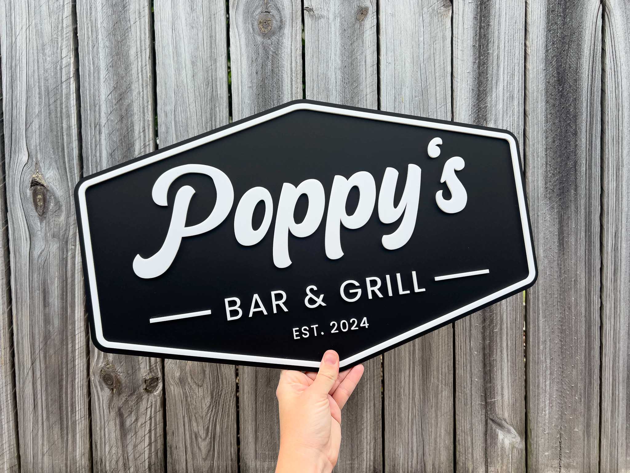 Hand holding a badge style signboard sign with 'Poppy's Bar & Grill' text against a wooden background