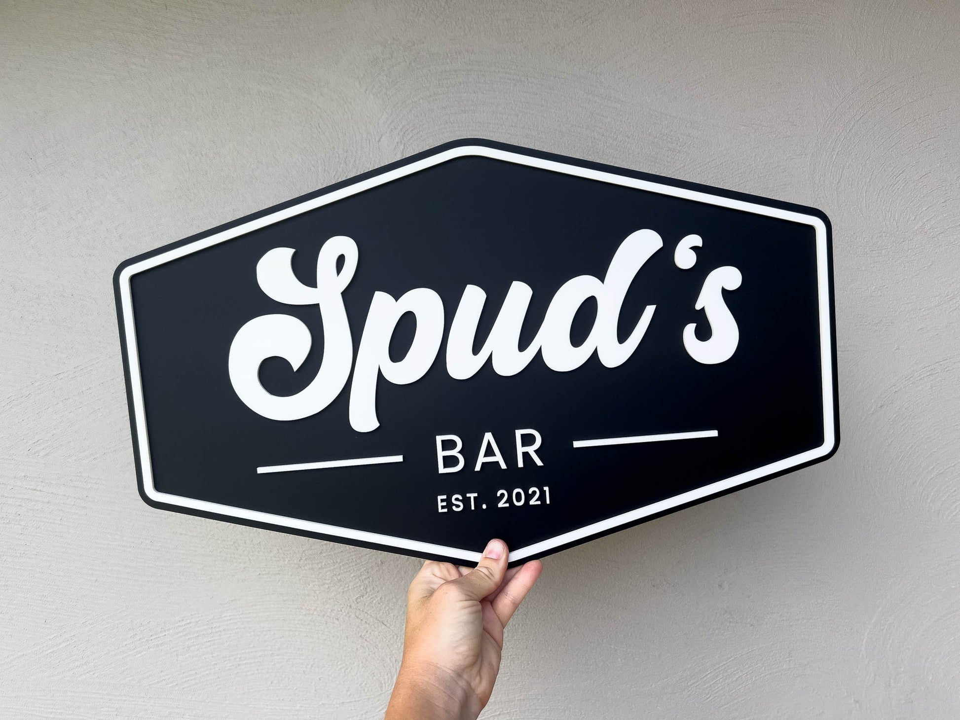 Hexagonal American badge style sign with 'Spud's Bar EST. 2021' text held against a plain background