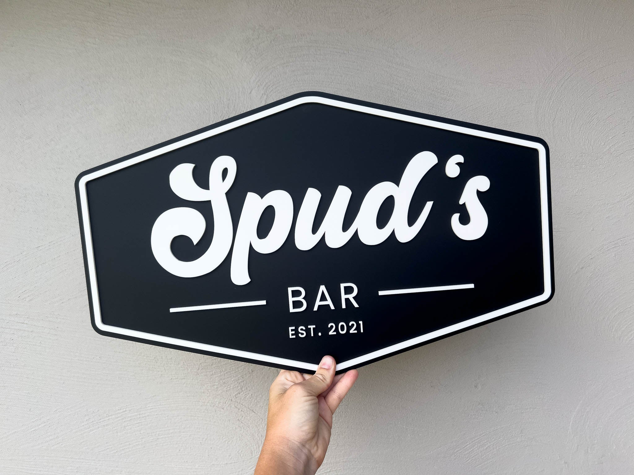 Hexagonal American badge style sign with 'Spud's Bar EST. 2021' text held against a plain background