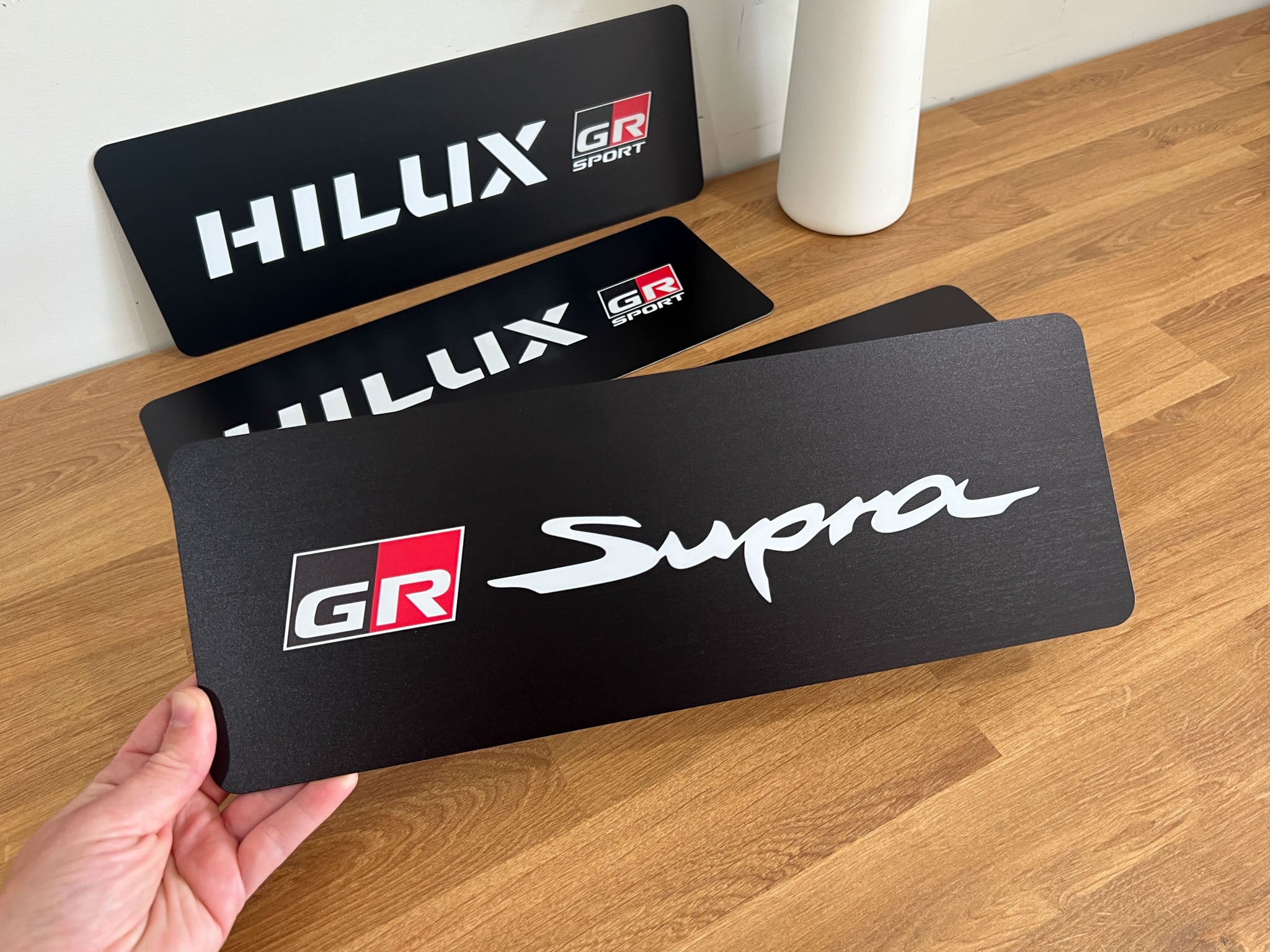 Black UV printed aluminium plates with 'HILUX', 'GR', and 'Supra' logos on a wooden surface.