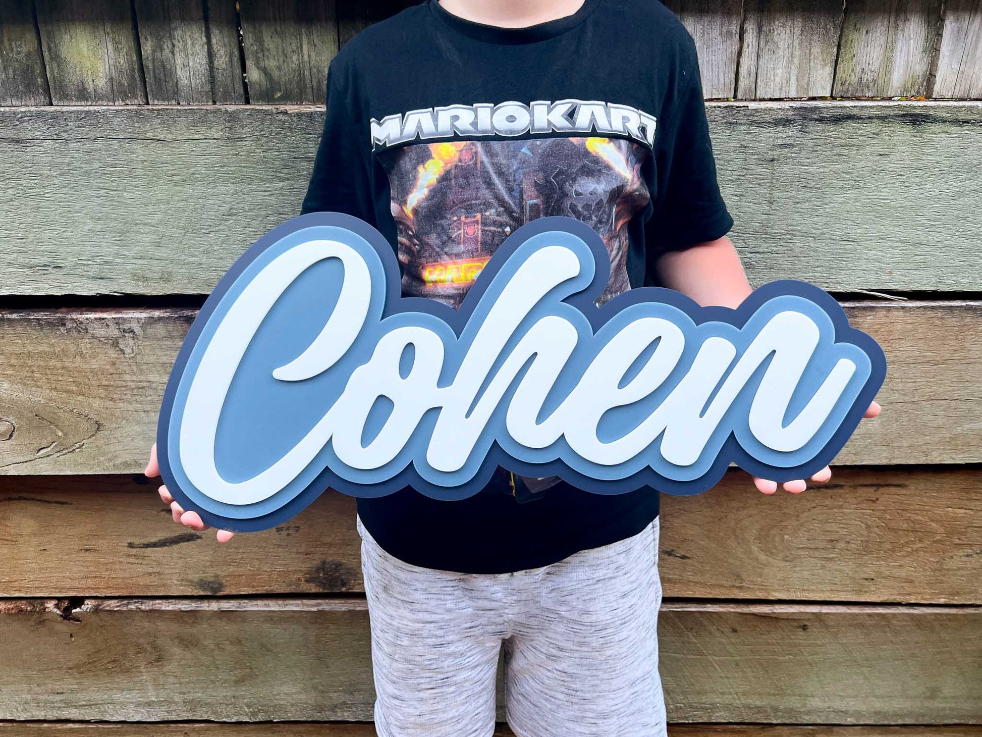 Child holding a large custom cut acrylic name plaque with the name 'Cohen' in shades of blue and white.