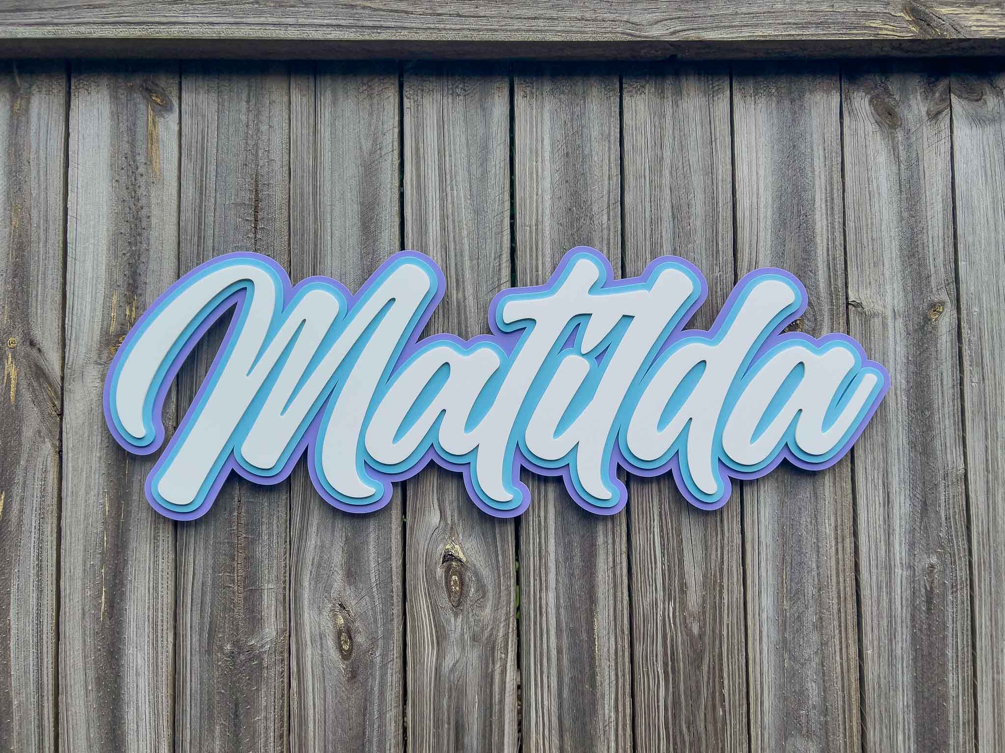 A surf typography style name plaque that says 'Matilda' that has been custom cut in purple, light blue and white acrylic.