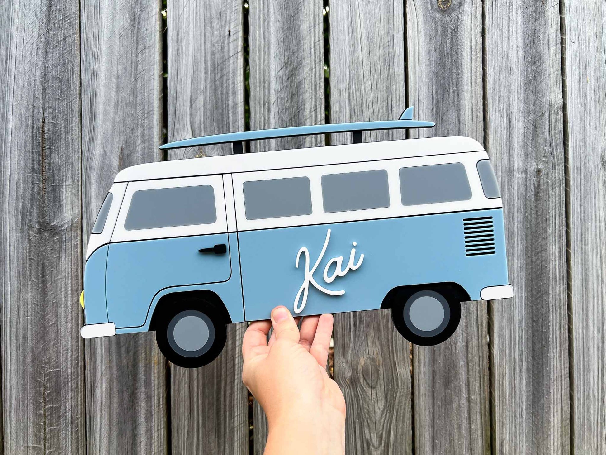 Hand holding a blue beach van-shaped sign with 'Kai' on a wooden background