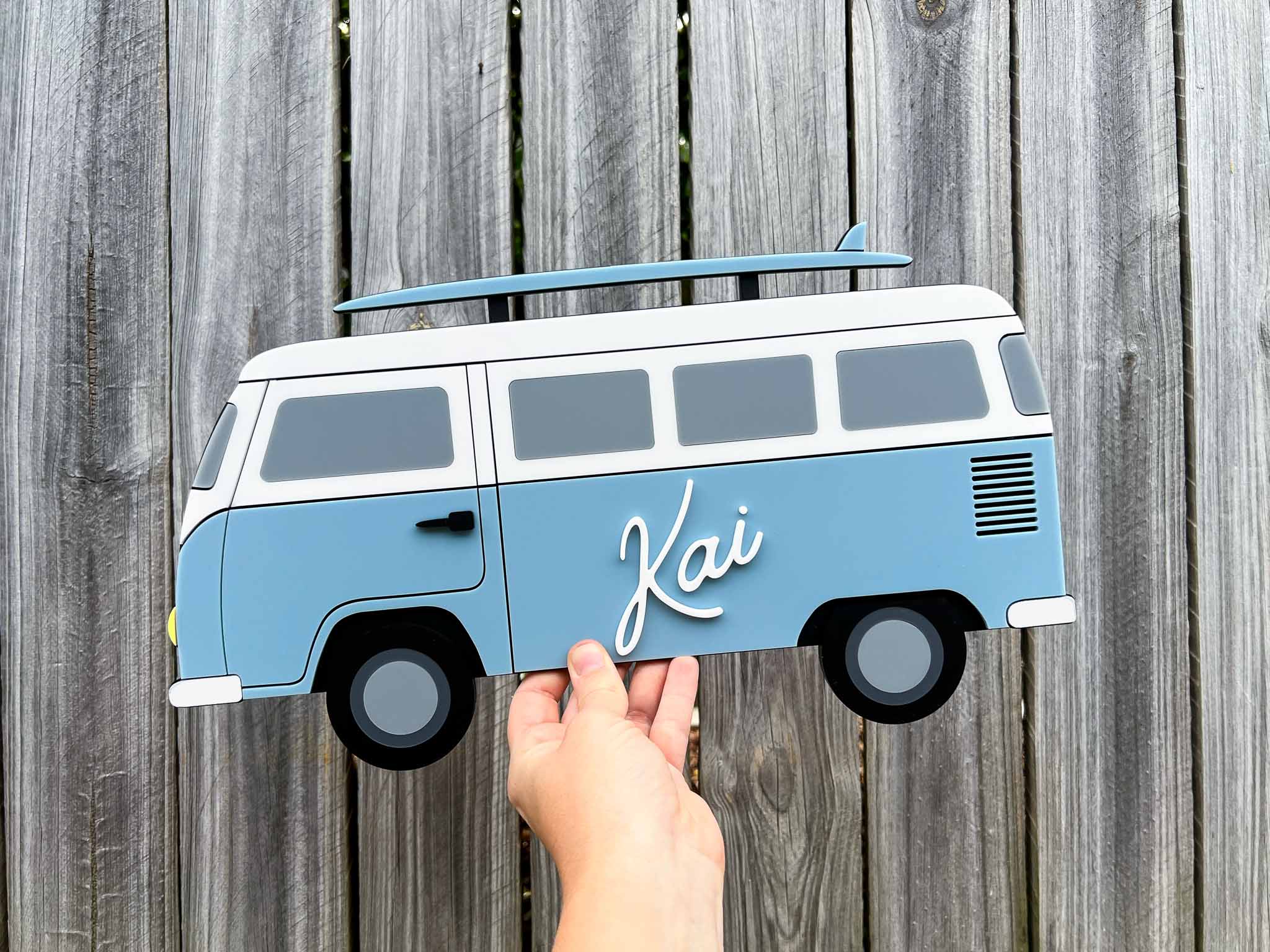 Hand holding a blue beach van-shaped sign with 'Kai' on a wooden background