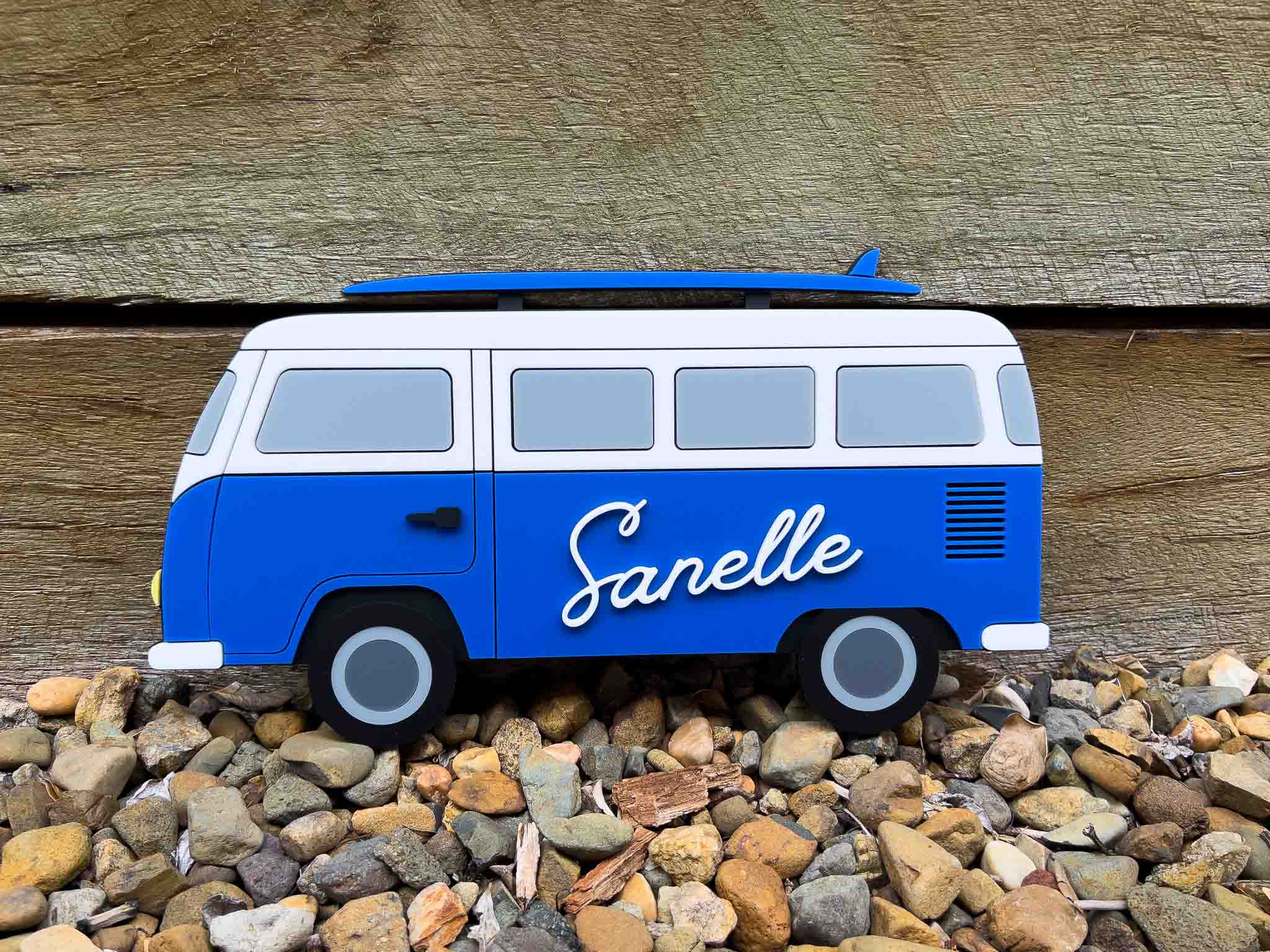 Blue and white van-shaped acrylic sign with the name 'Sanelle' on a pebble surface.