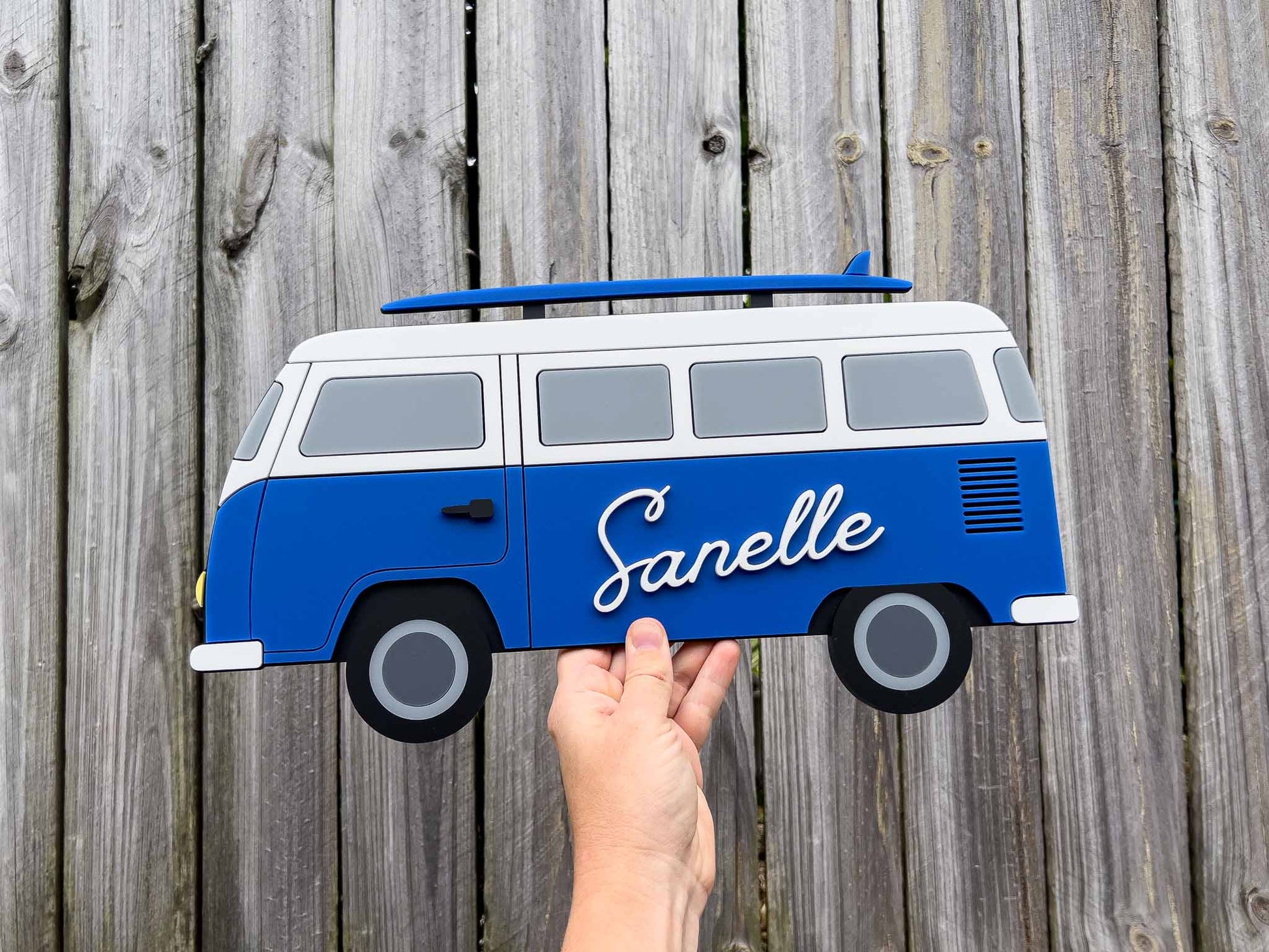 Hand holding a custom made 3D blue and white surf van-shaped sign with 'Sanelle' on it against a wooden background