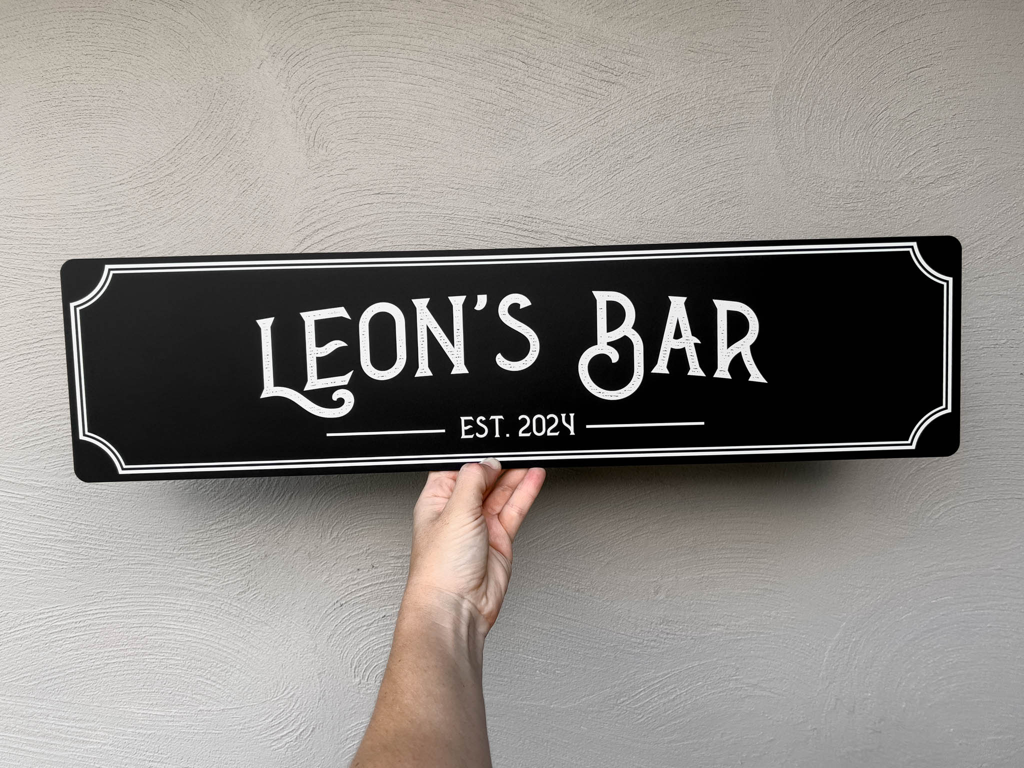 Hand holding a black sign with 'LEON'S BAR EST. 2024' on a light gray background