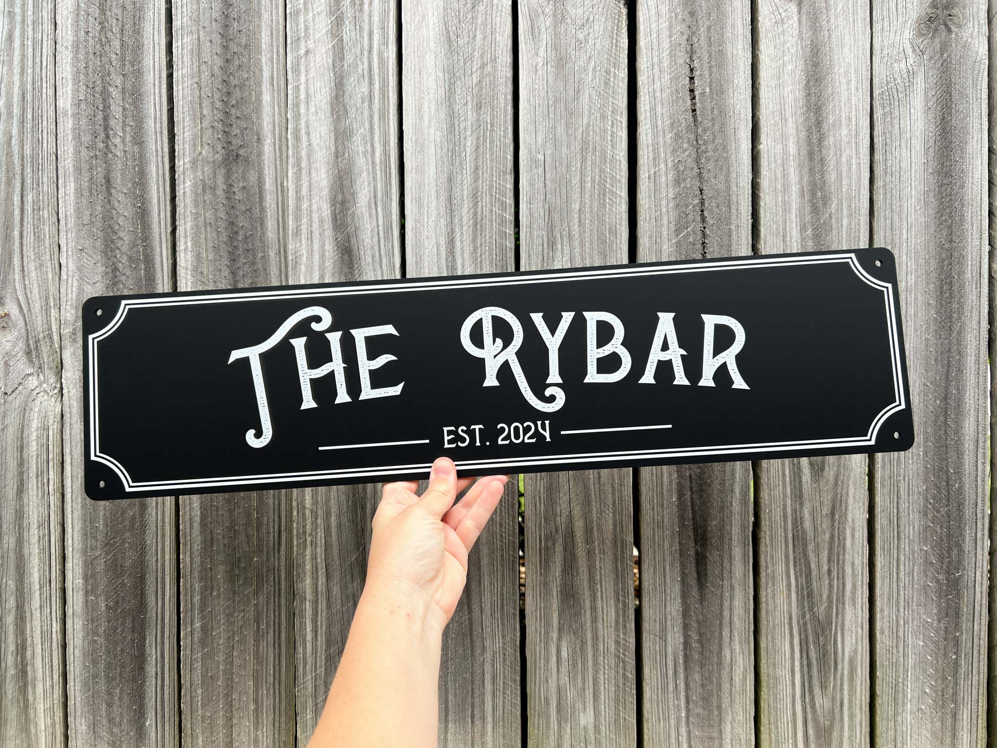 Hand holding a custom printed acrylic sign with 'The Rybar' and 'Est. 2024' against a wooden background